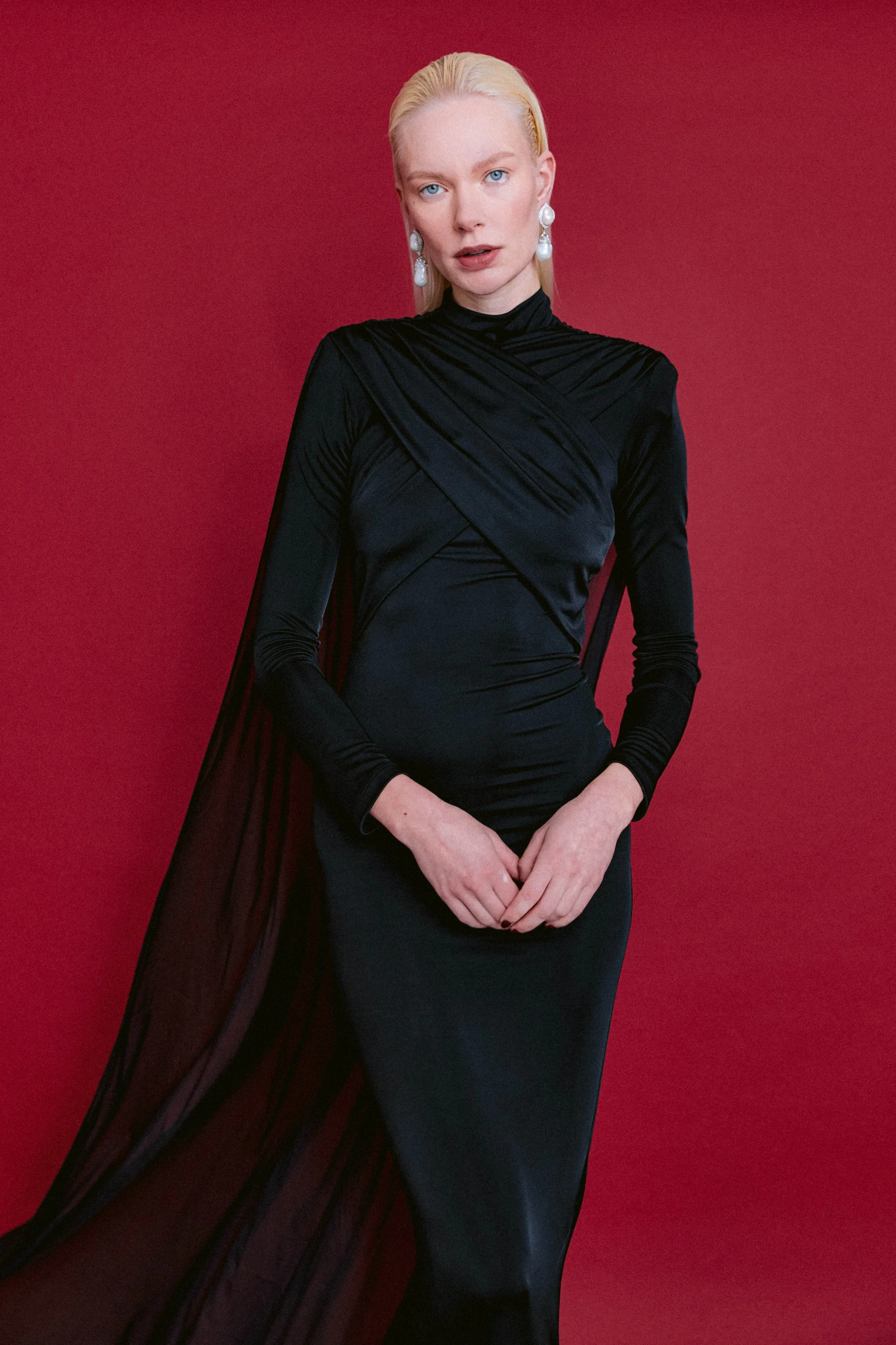 A woman with platinum blonde hair, blue eyes, and fair skin wearing a black, long-sleeved, fitted dress with a cape, standing against a deep red background.