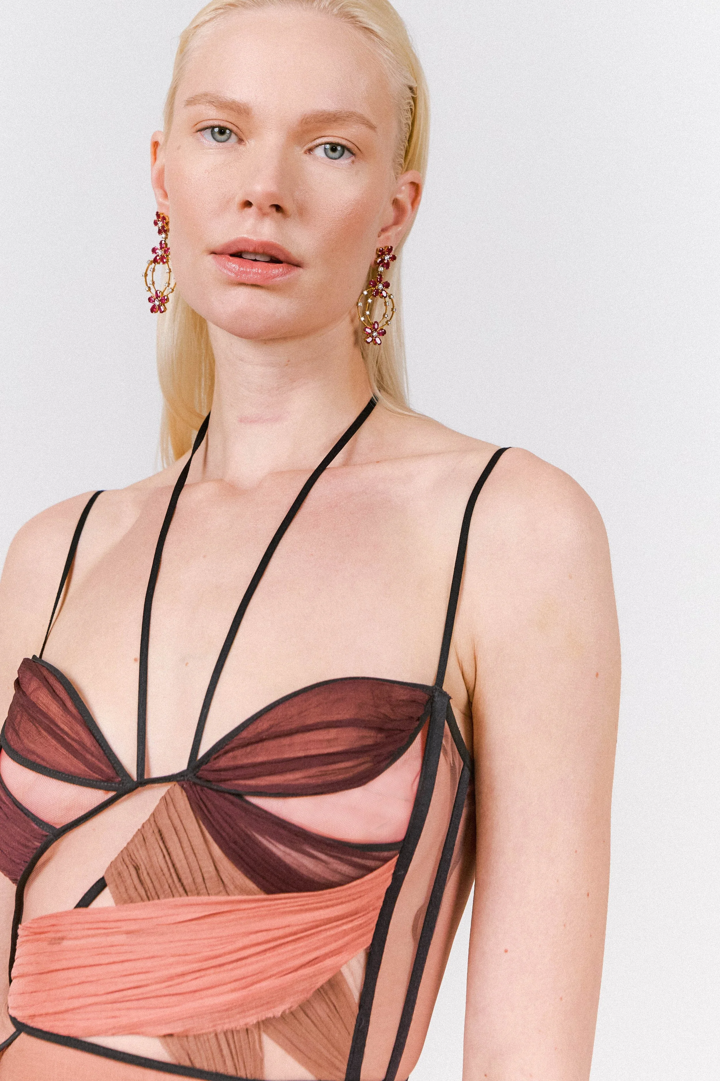 A woman with blonde hair, blue eyes, and light makeup wearing colorful earrings and a multicolored, strappy, sheer top.