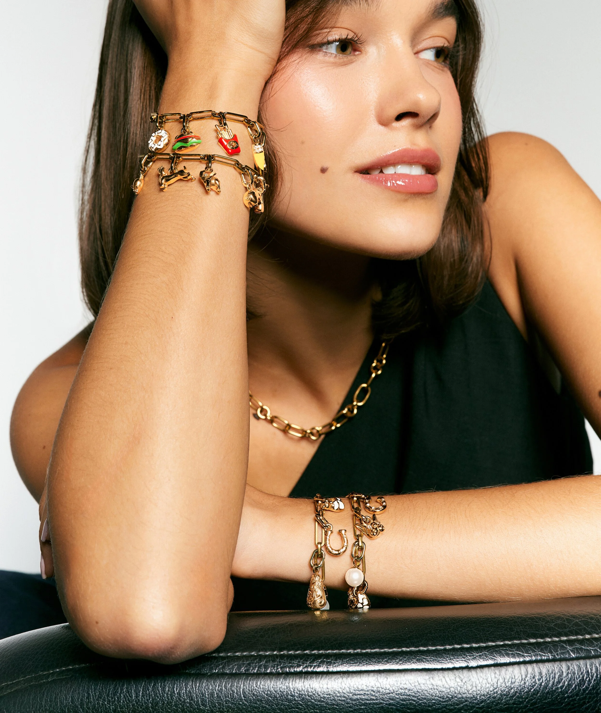 Close-up of a woman wearing multiple gold and charm jewelry bracelets, chains, and charms on her wrist and neck, resting her head on her hand with a slight smile.