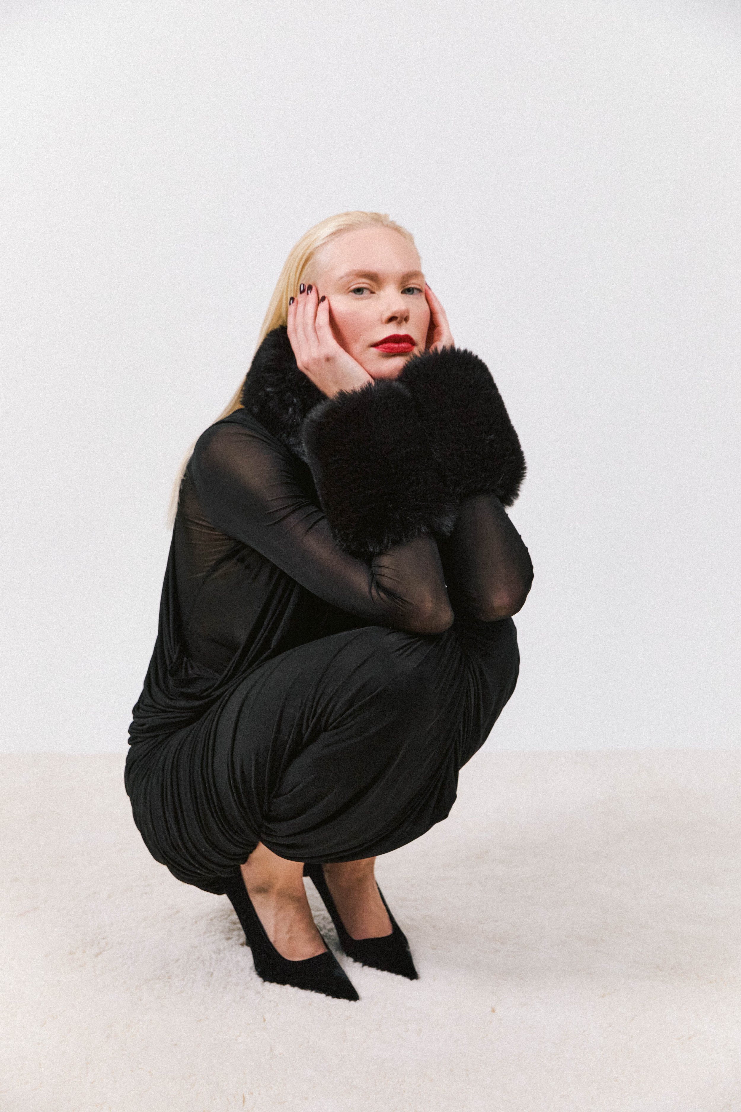 Woman in black outfit with fur cuffs squatting on white carpet, looking at the camera, against a plain white background.