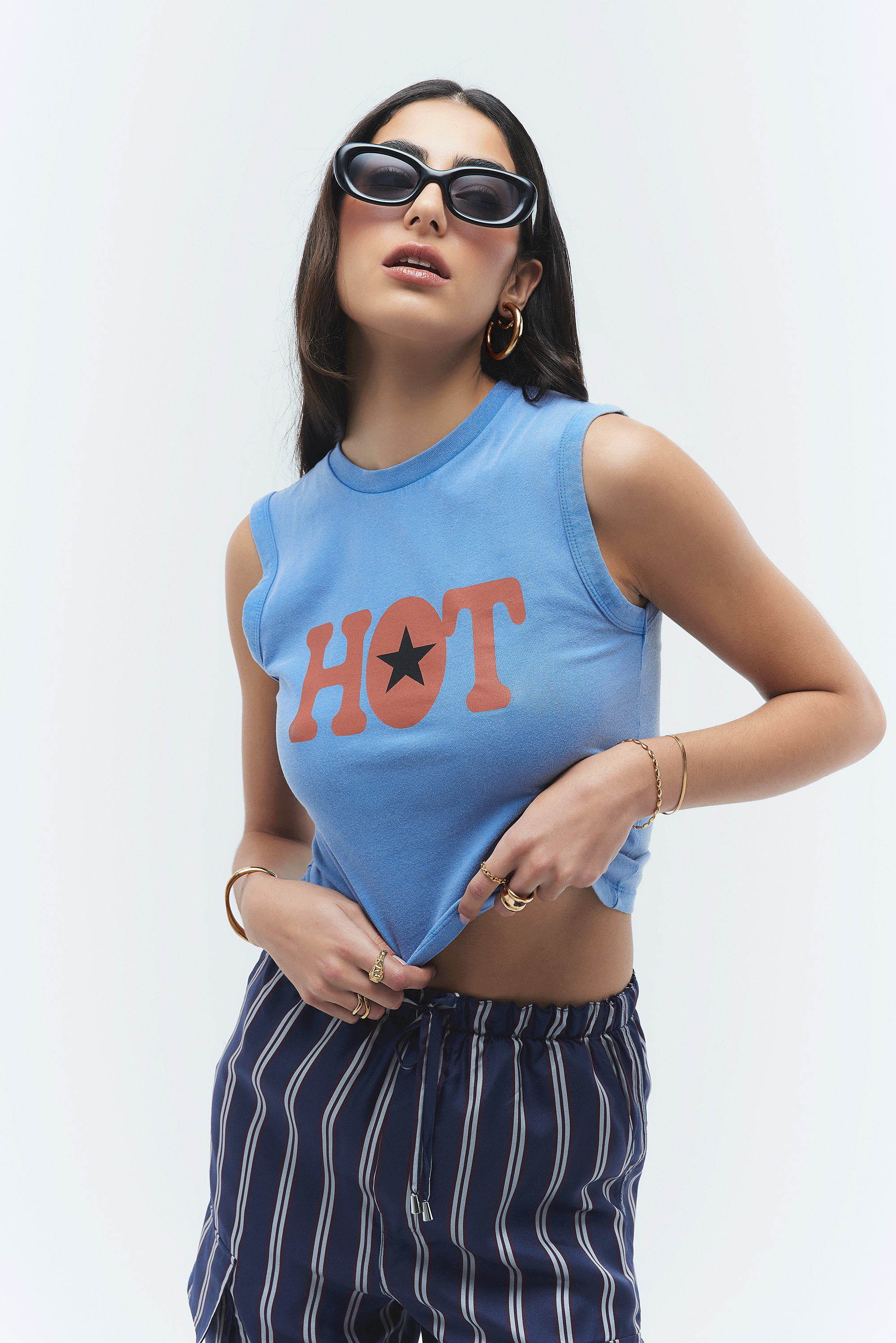 Woman wearing black sunglasses, gold hoop earrings, and multiple gold rings and bracelets, lifting her cropped blue tank top with 'HOT' written on it, against a plain white background.