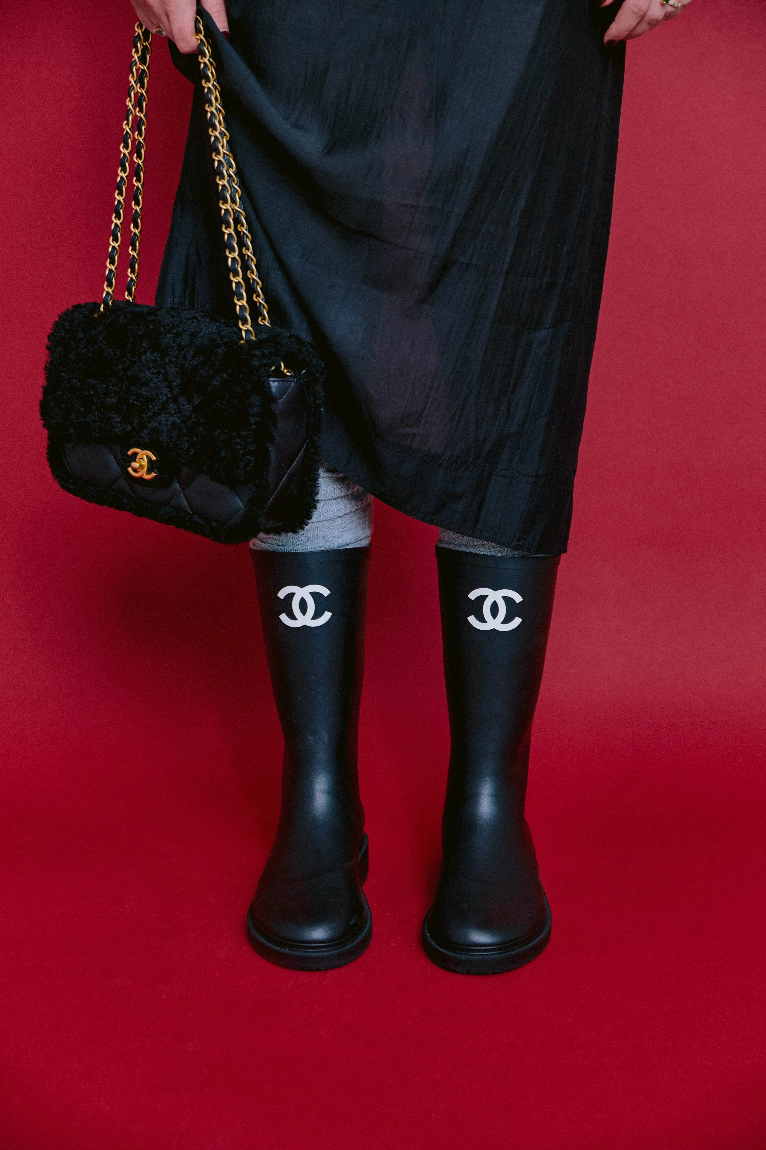 Person wearing black rain boots with white Chanel logo, holding a black Chanel purse, standing against a red background.