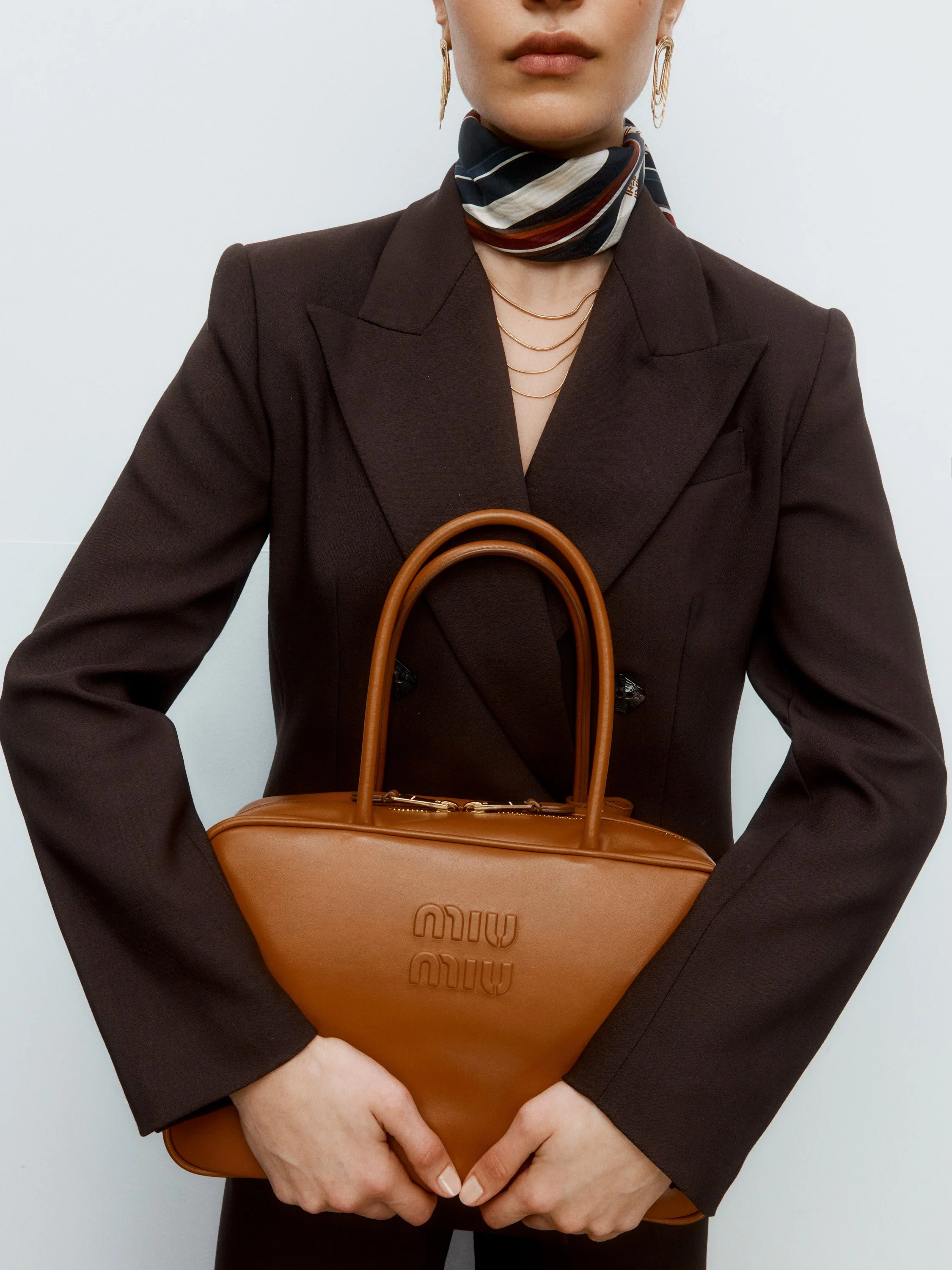 A woman wearing a black blazer, striped scarf, and gold jewelry holding a tan Miu Miu handbag.