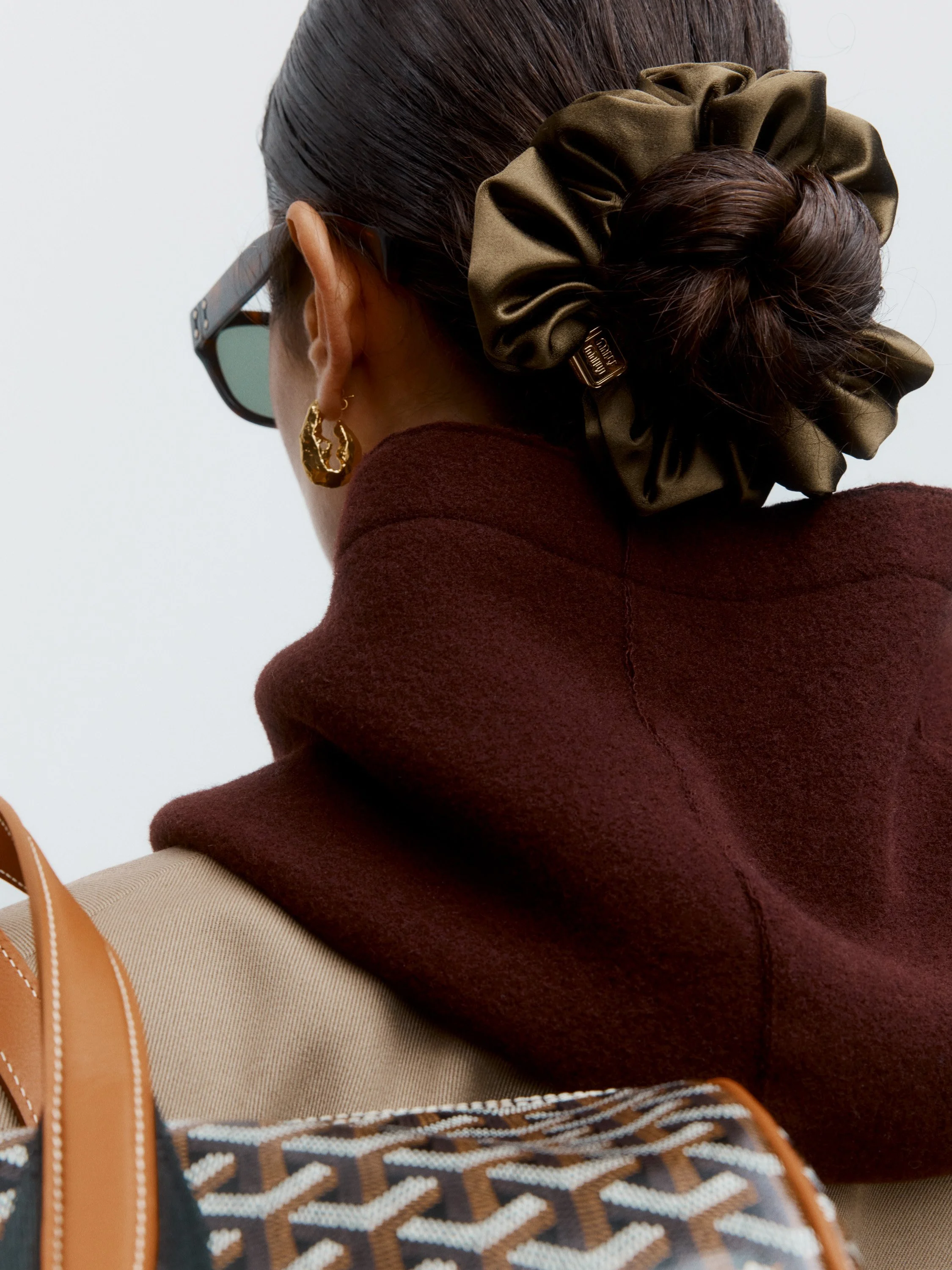 A woman with dark brown hair styled in a bun with a large olive-green satin scrunchie, wearing gold hoop earrings, dark sunglasses, a burgundy turtleneck, and carrying a patterned bag with tan straps.