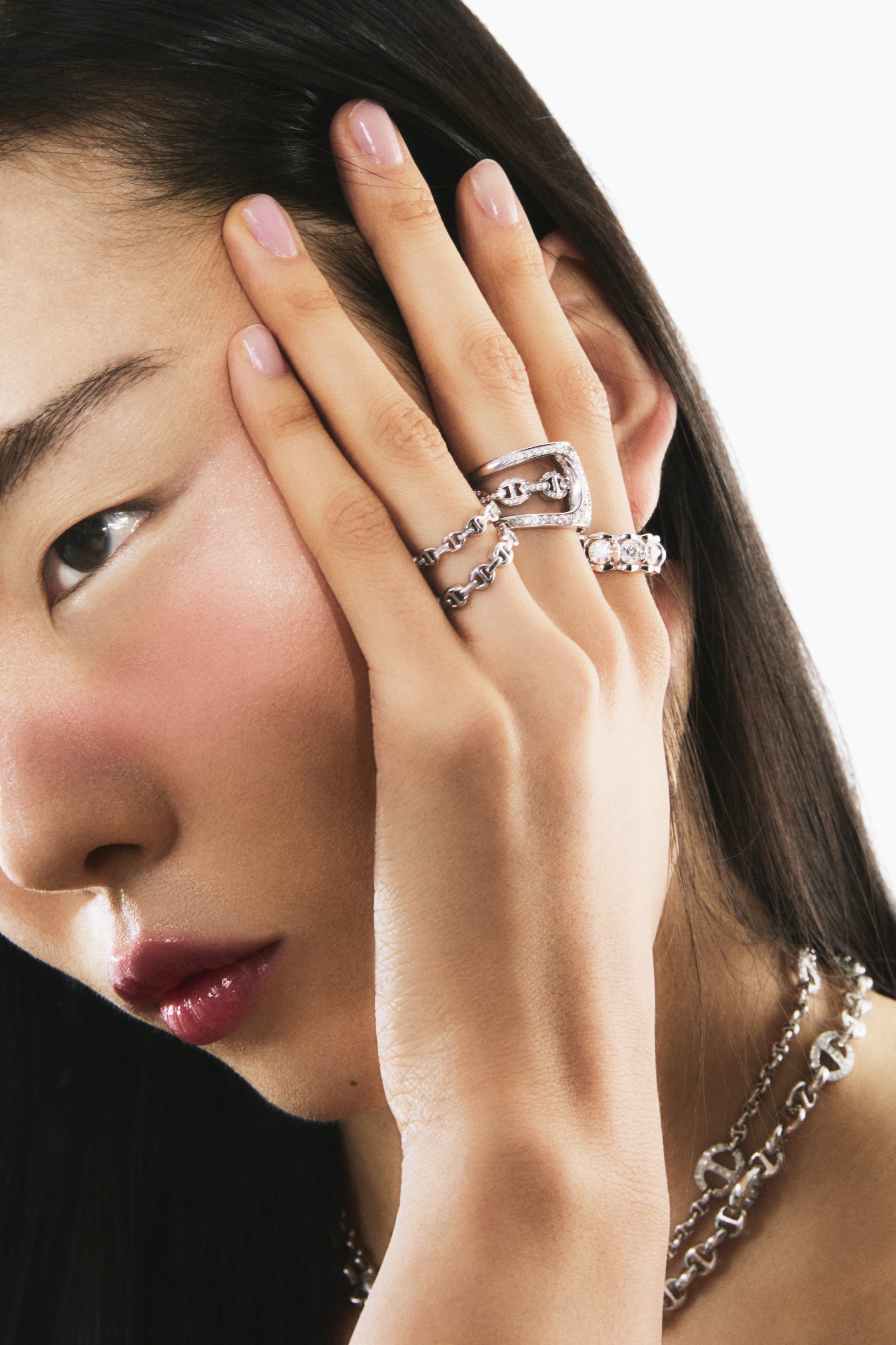 Close-up of a woman with long dark hair, wearing multiple silver jewelry rings and a necklace, resting her hand on her face with her eyes partially closed.