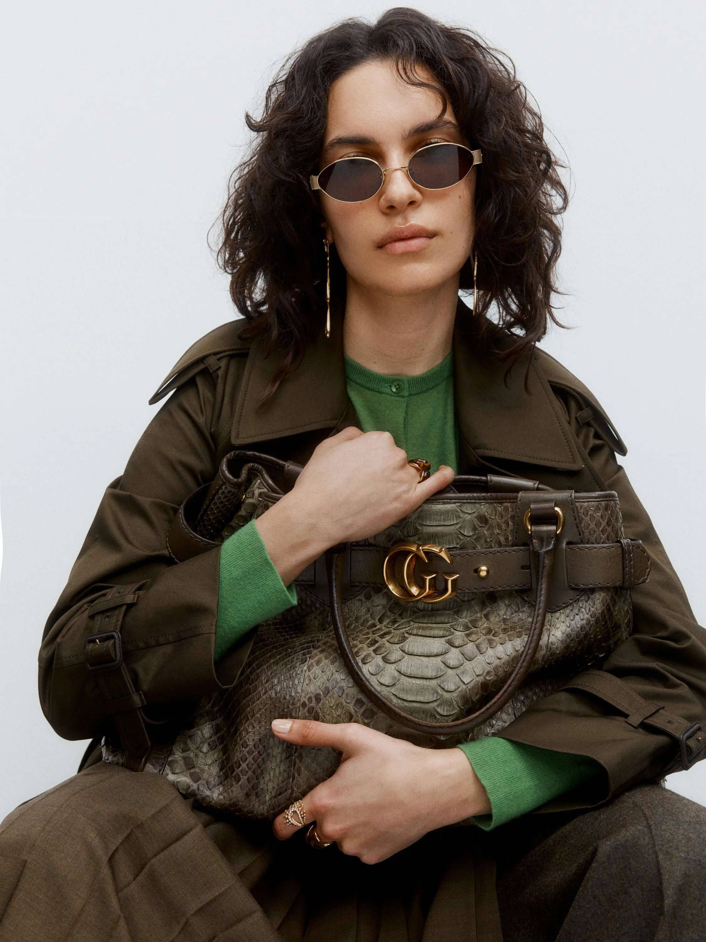Fashion model wearing sunglasses, a green sweater, a khaki trench coat, and holding a snakeskin-patterned handbag with gold GG Gucci logo.