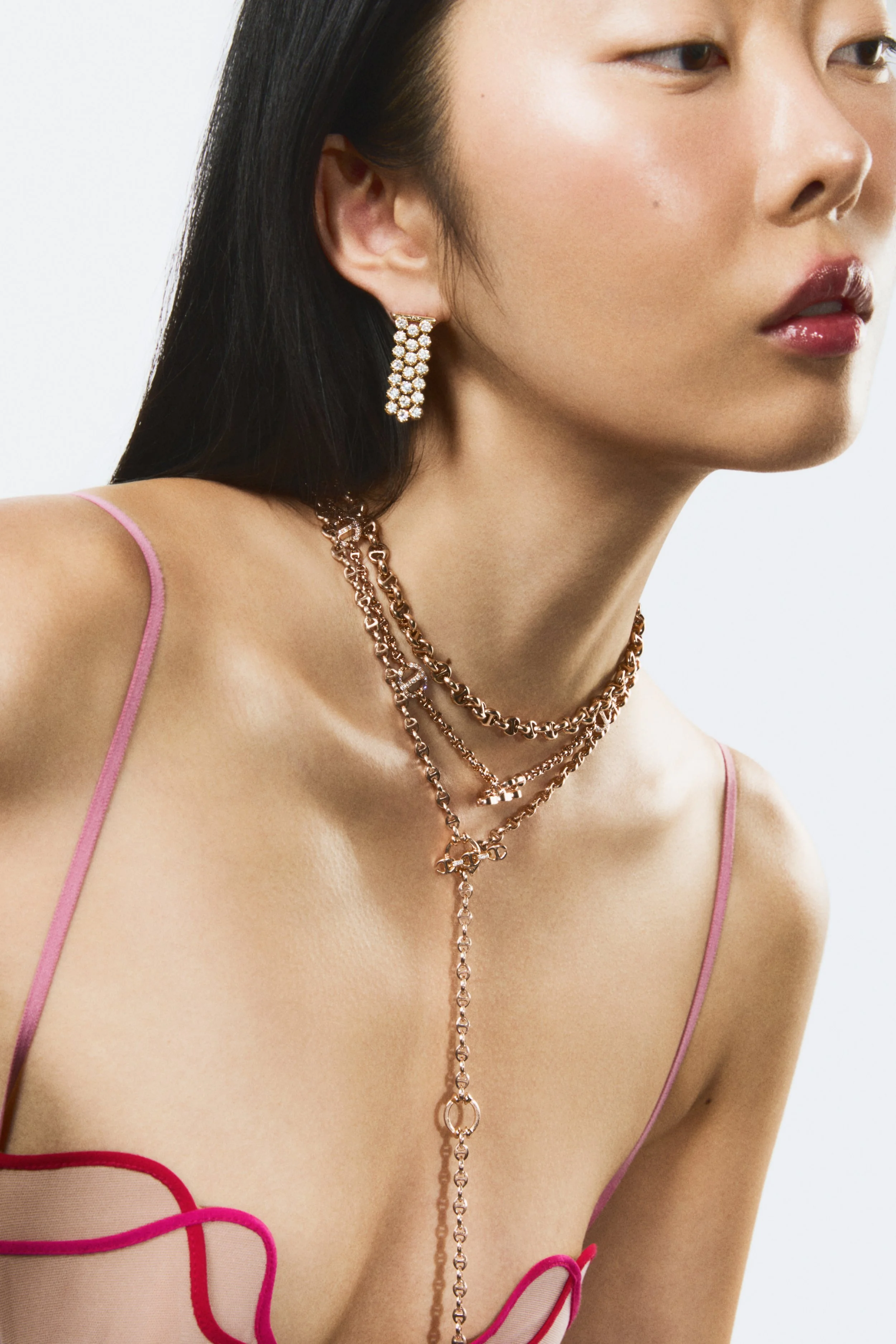 A close-up of a woman wearing rose gold jewellery, including layered necklaces and sparkling earrings, with a pink strappy top.