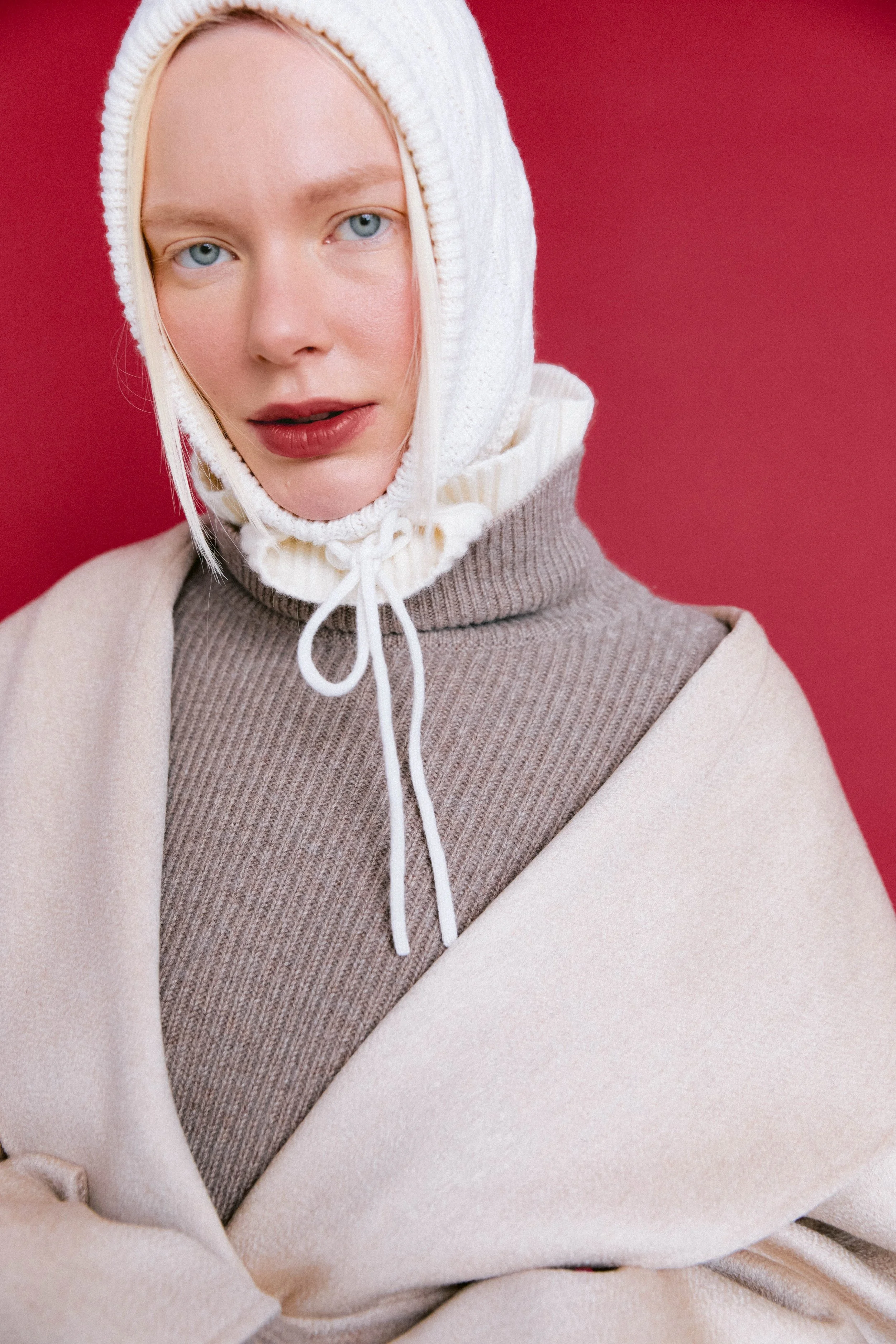 A woman with blue eyes wearing a white knit hood and a beige and taupe layered outfit against a red background.