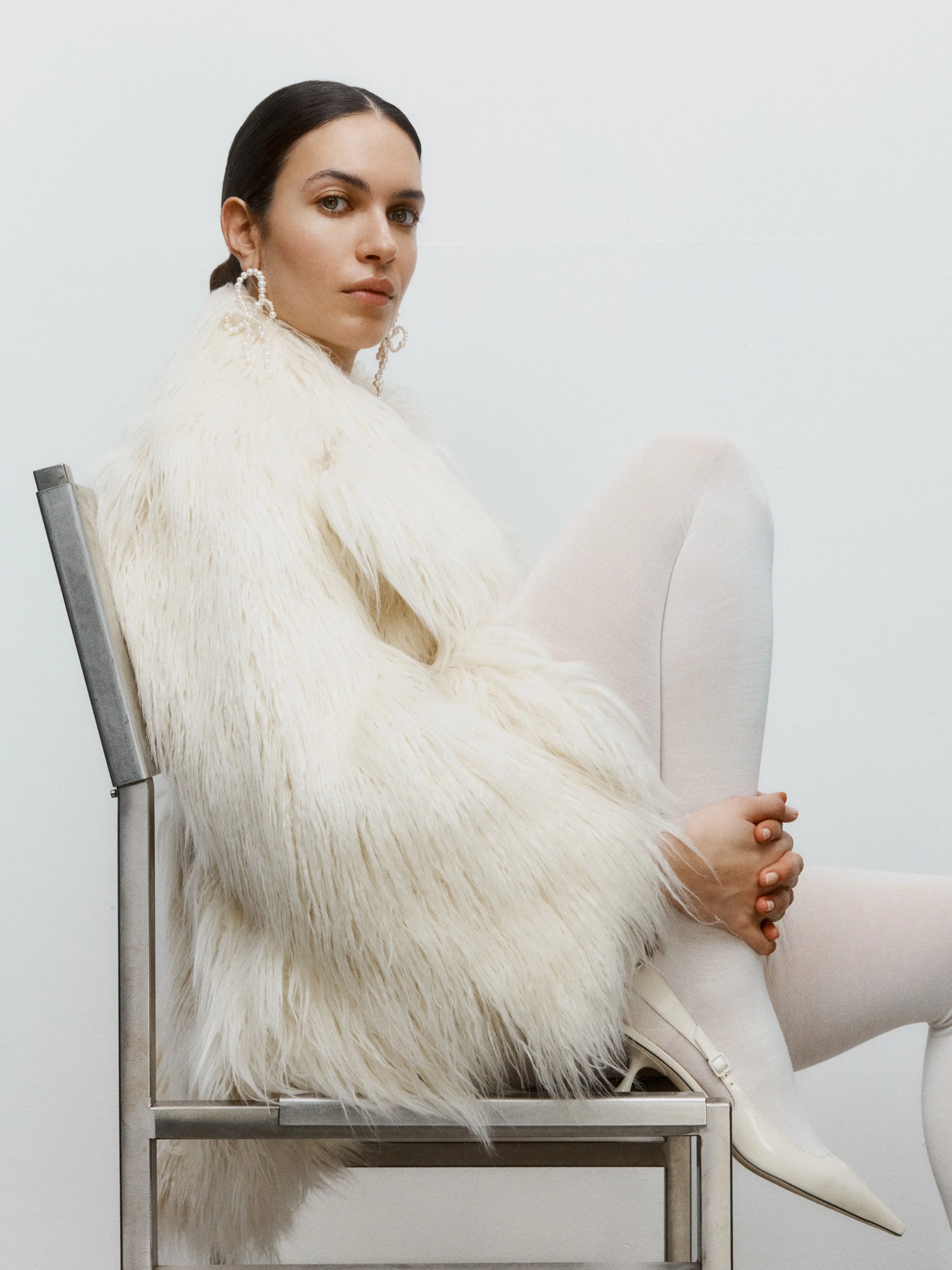 A woman with dark hair, wearing large pearl earrings, sits sideways on a metallic chair. She is dressed in a white fur coat, white tights, and white pointed-toe heels, against a plain white background.