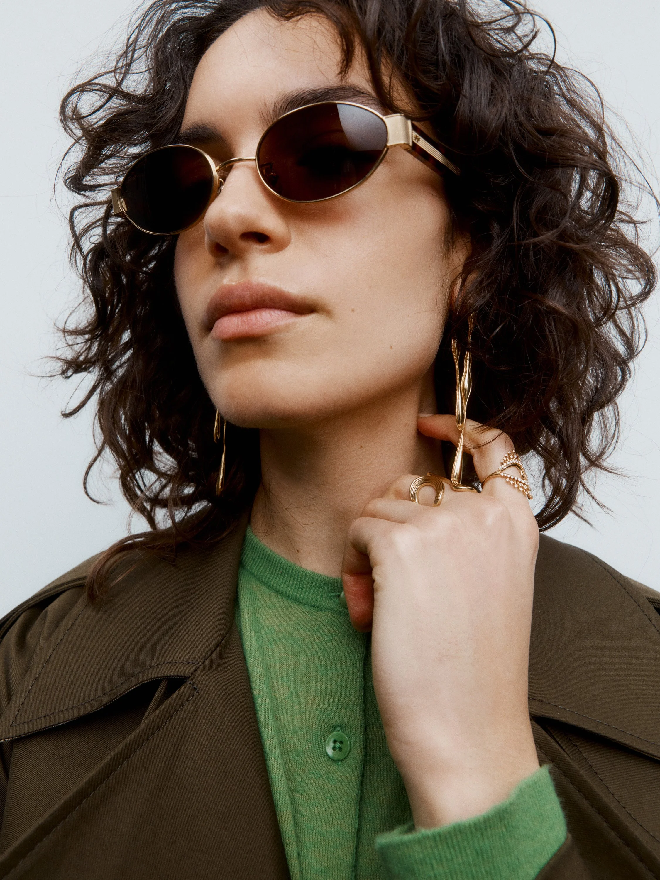 Close-up of a woman wearing oversized sunglasses with dark lenses, gold jewelry including earrings and rings, a green sweater, and a brown jacket, with her hand resting near her face.
