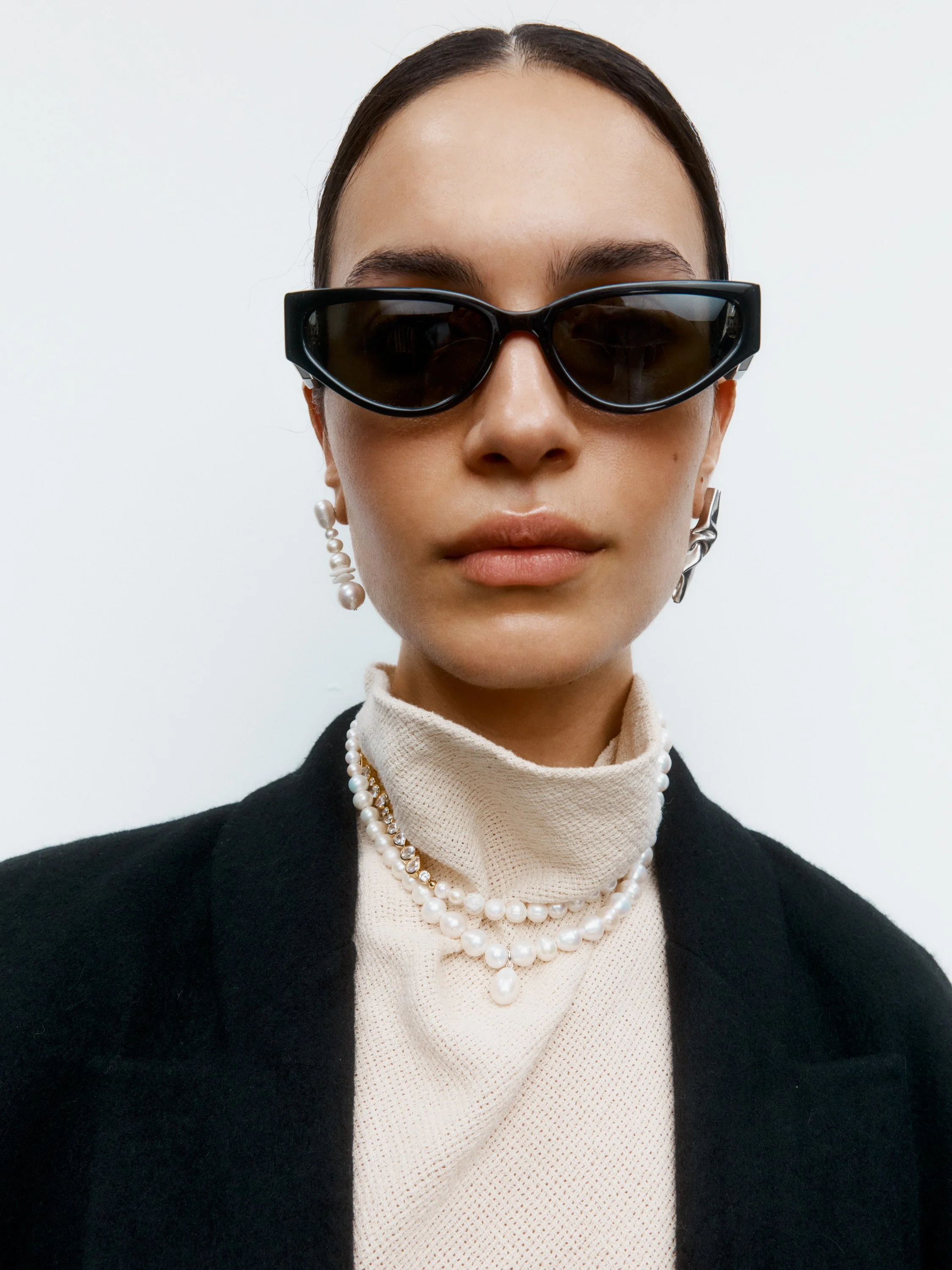 A woman with dark hair wearing black sunglasses, pearl earrings, a layered pearl necklace, a cream turtleneck, and a black blazer standing against a plain white background.