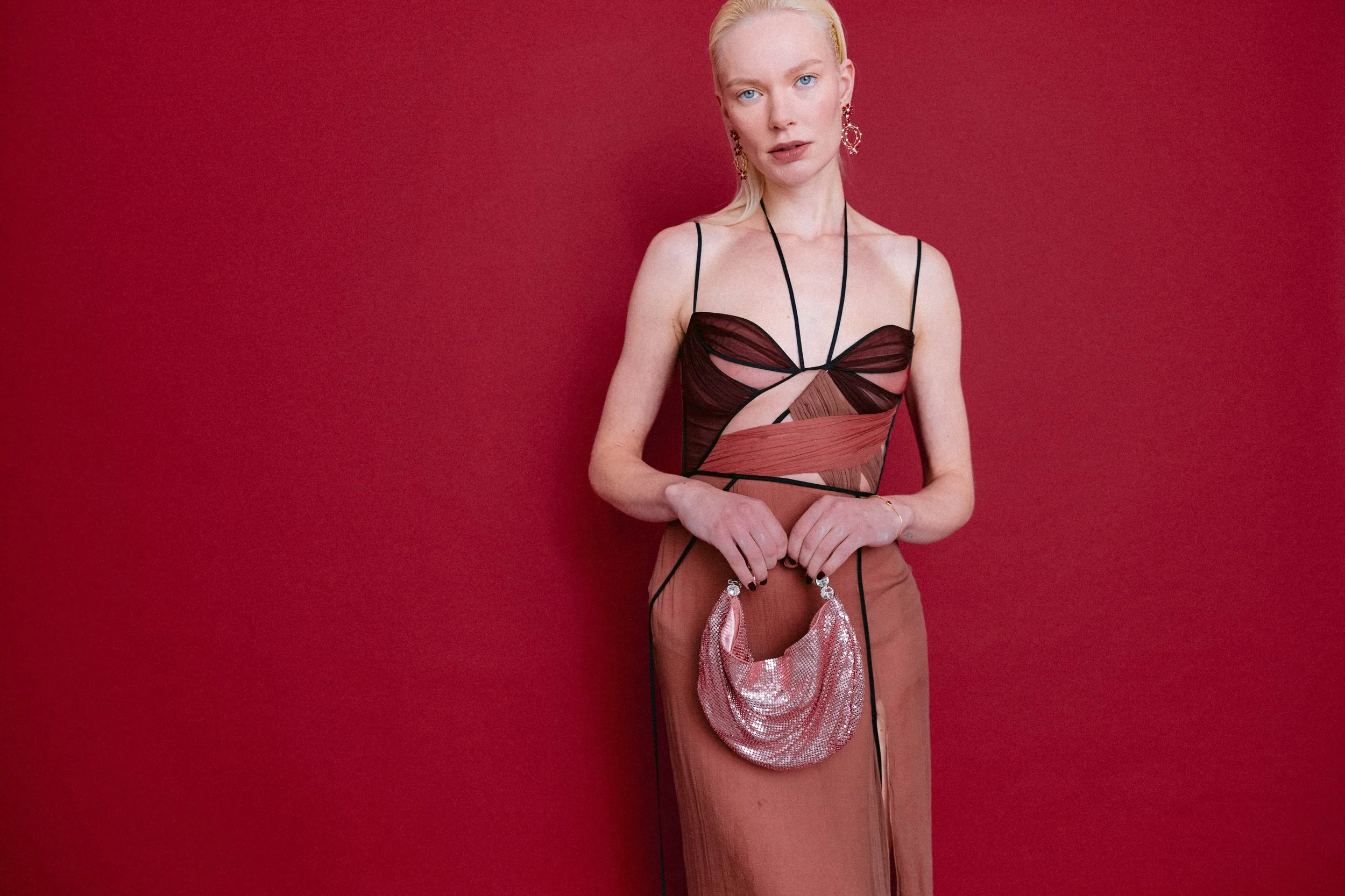 Blonde woman wearing a black and pink tone cut-out dress with a pink sequin handbag, standing against a red background.