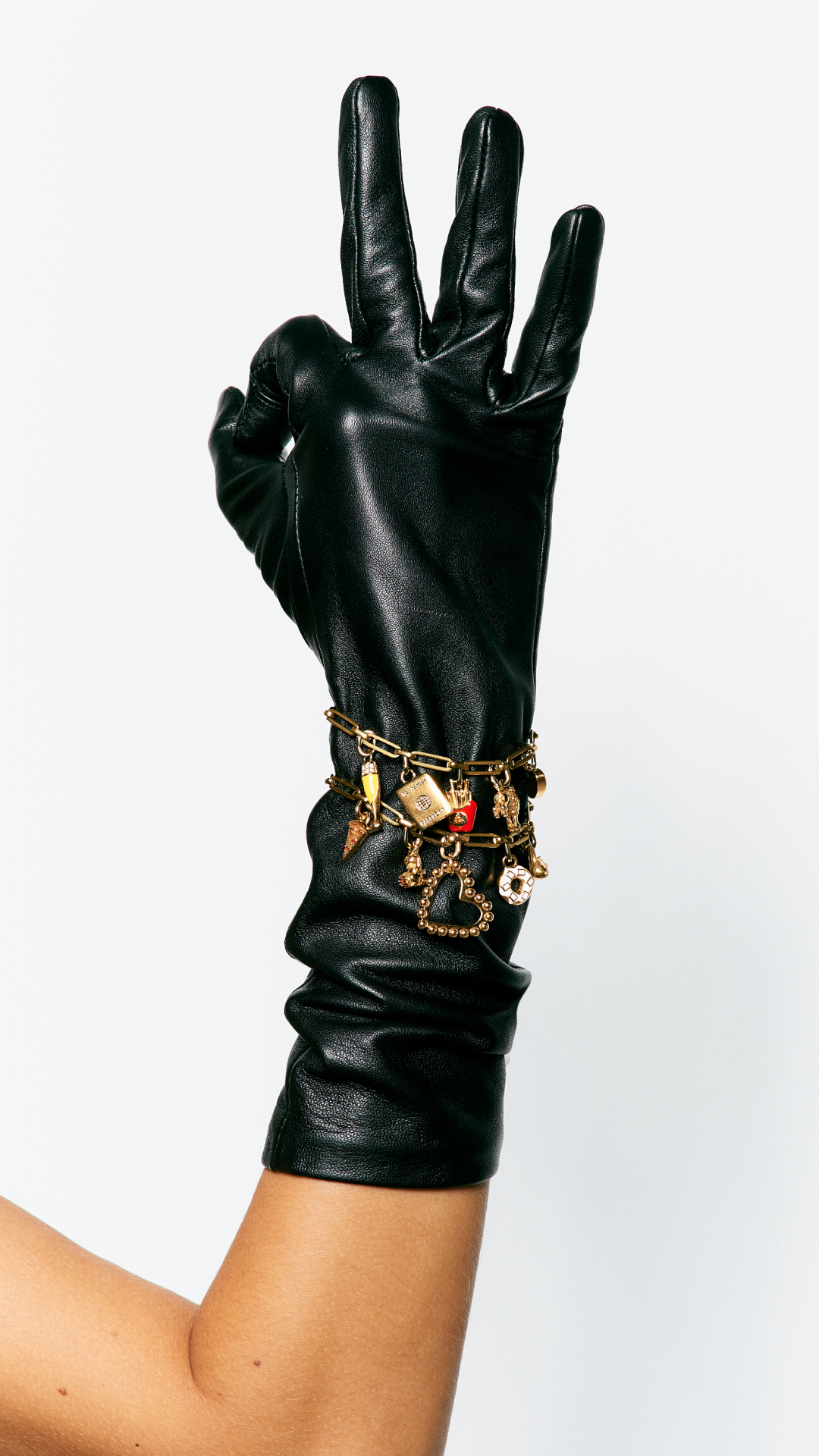 A black leather glove adorned with gold charm bracelets and rings, with a person's arm visible.