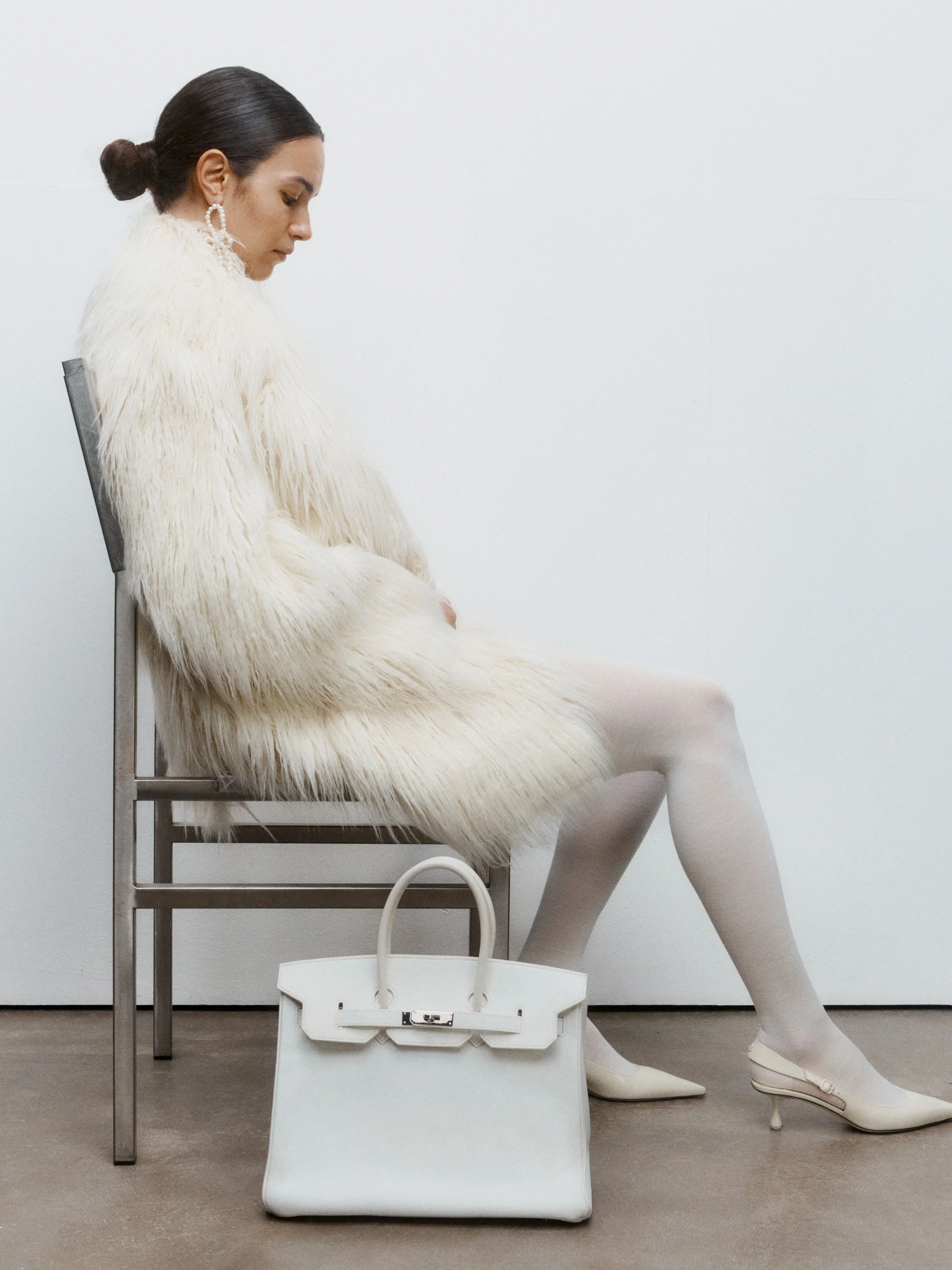A woman with dark hair in a bun, wearing a white faux fur coat, white tights, and white heels, sits on a metal chair. There is a white handbag on the floor beside her and a plain white wall in the background and concrete floor.