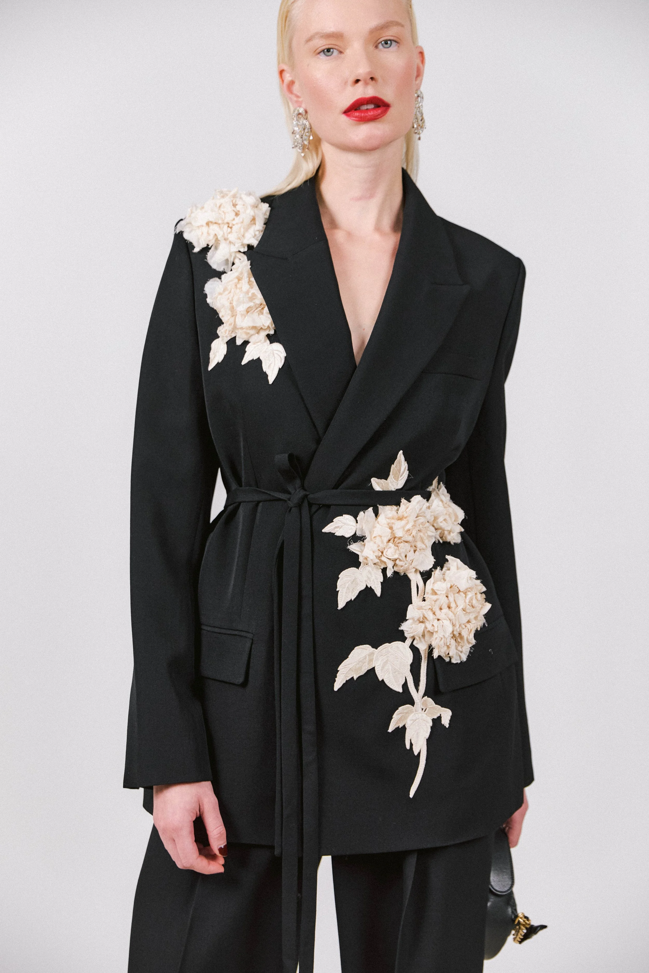 A woman wearing a black blazer with beige floral embroidery, red lipstick, and large earrings, posing against a plain background.