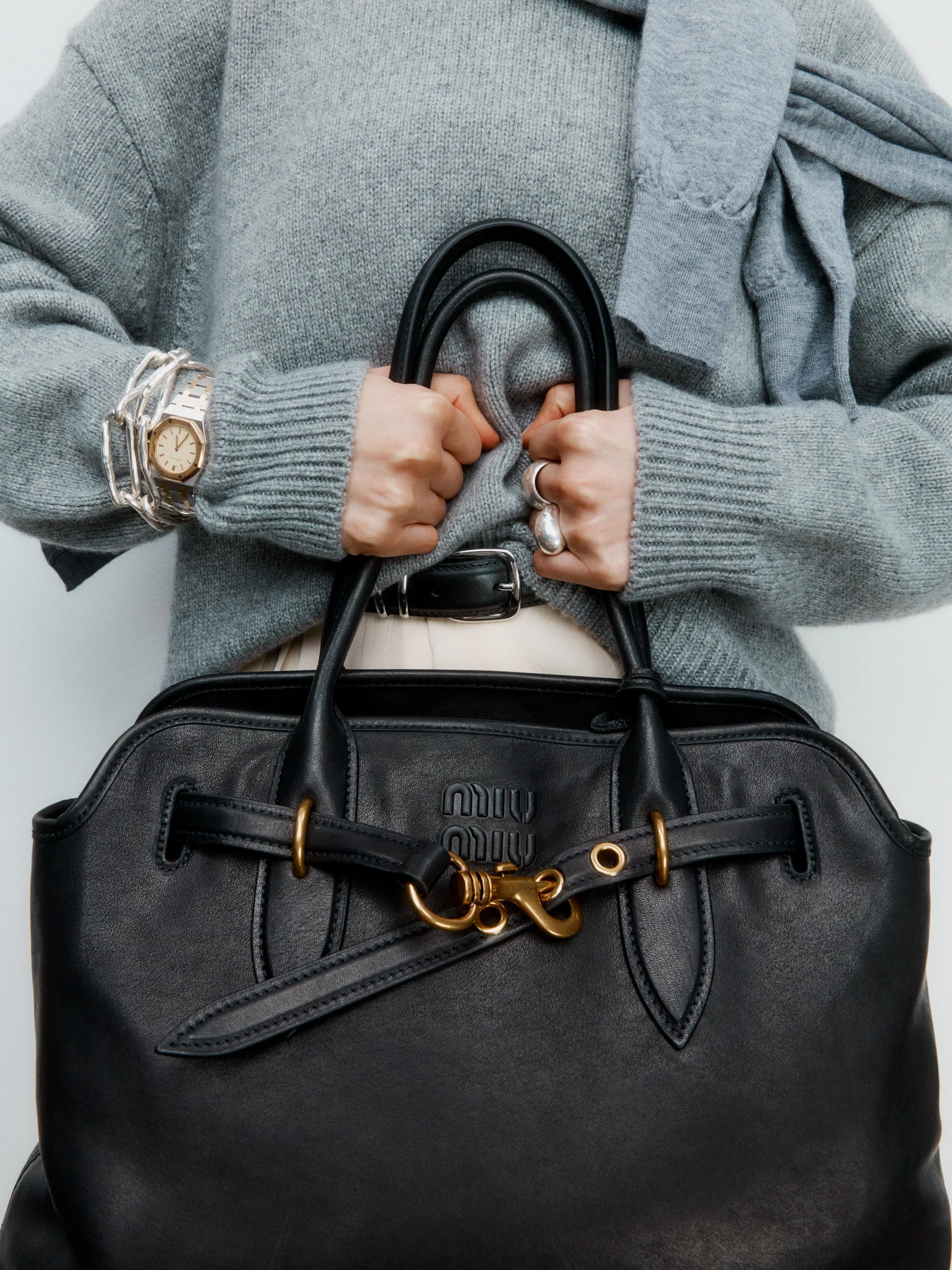 Person holding a black Miu Miu handbag, wearing a gray sweater, with a gold and silver watch and silver rings.