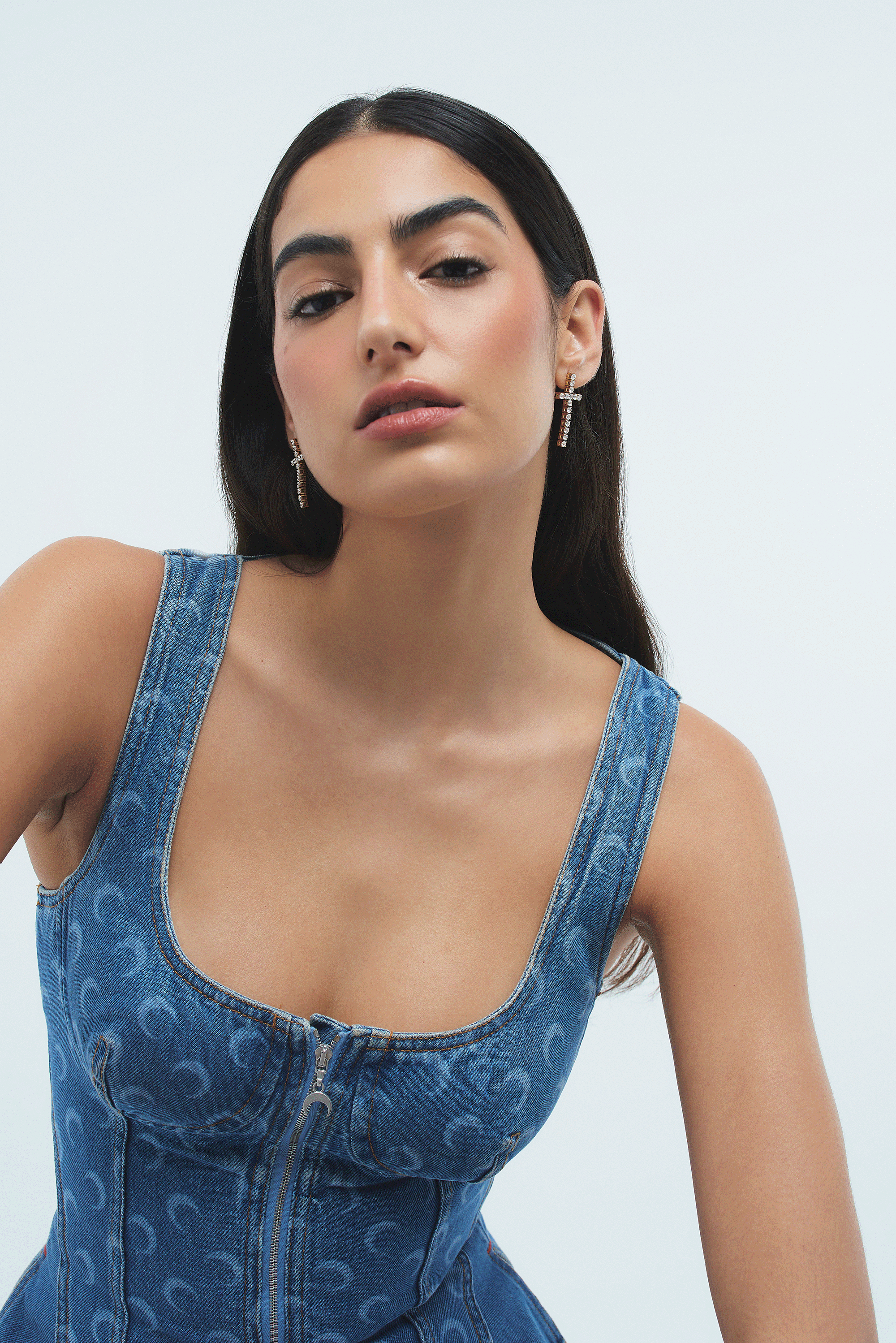 A woman with long dark hair wearing a sleeveless denim dress with a zip in the front and decorative patterns, along with dangling earrings, posed against a plain background.