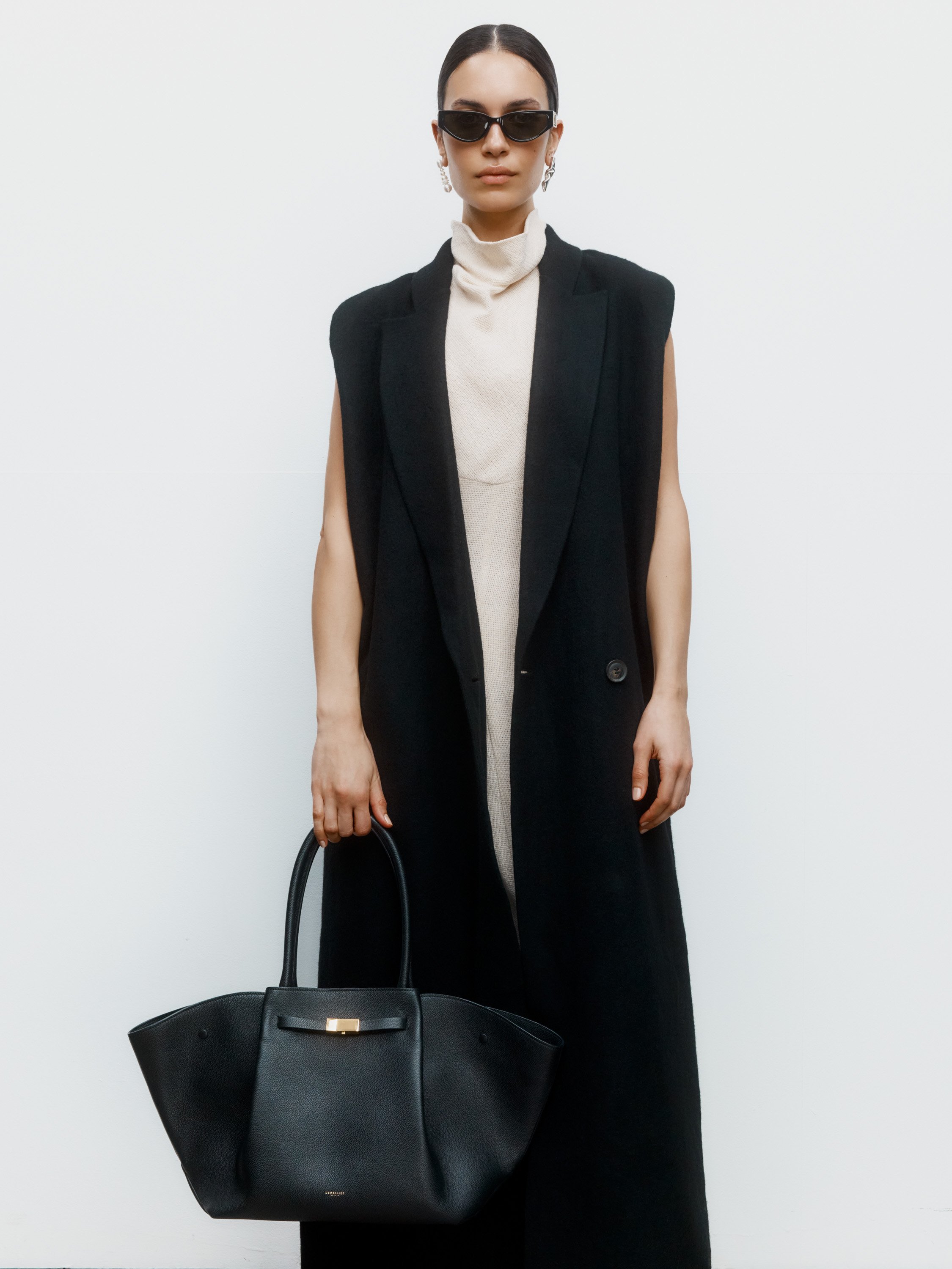 Stylish woman wearing black sunglasses, earrings, a beige turtleneck dress, a black sleeveless blazer, and holding a large black handbag, standing against a white background.