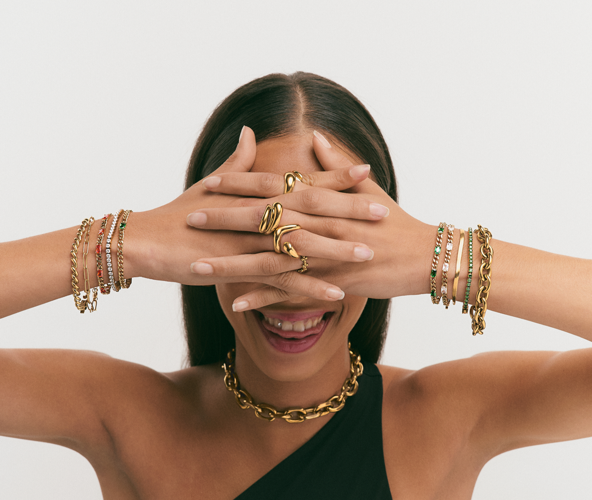 A woman with dark hair, smiling with her mouth open, wearing multiple gold bracelets, rings, and a gold chain necklace. She covers her eyes with her hands, which are also adorned with rings and bracelets. She is wearing a one-shoulder black top.