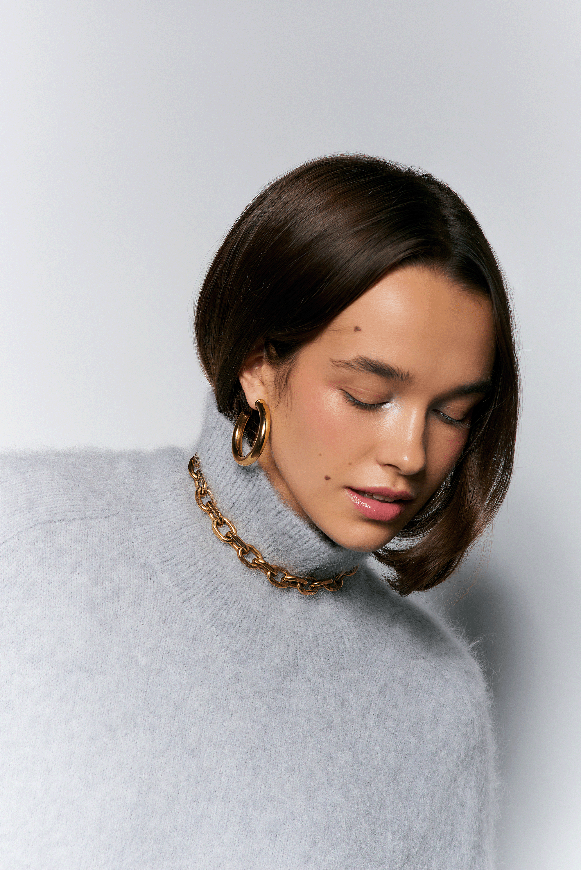 A woman with short brown hair wearing a gray turtleneck sweater, gold hoop earrings, and a gold chain necklace, with her eyes closed.