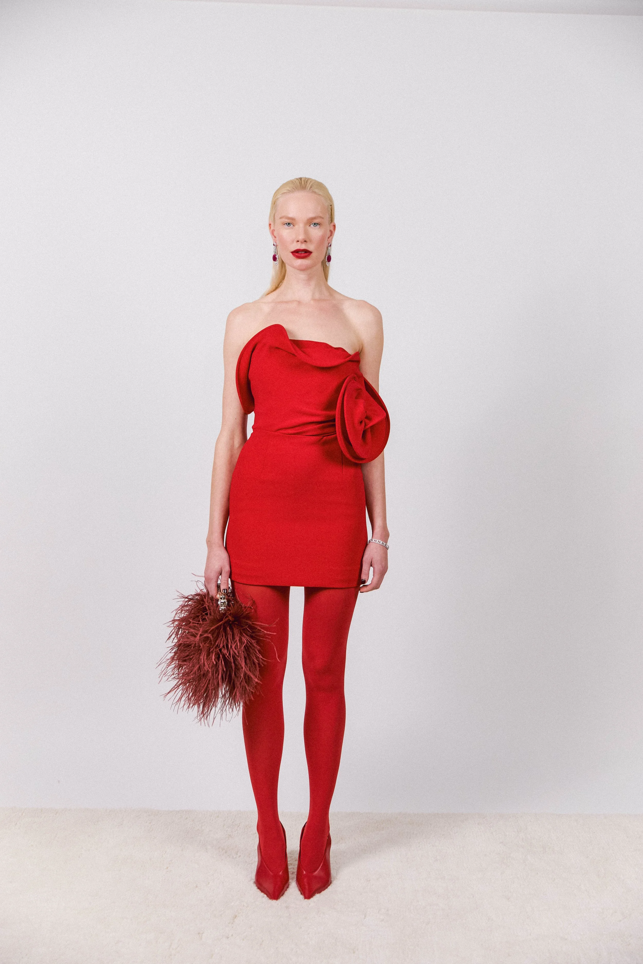 A woman in a red strapless dress with draped fabric and a large fabric flower embellishment on the front, matching red tights, red high heels, holding a brown feathered clutch, with blonde hair and pink earrings, standing against a plain white background.
