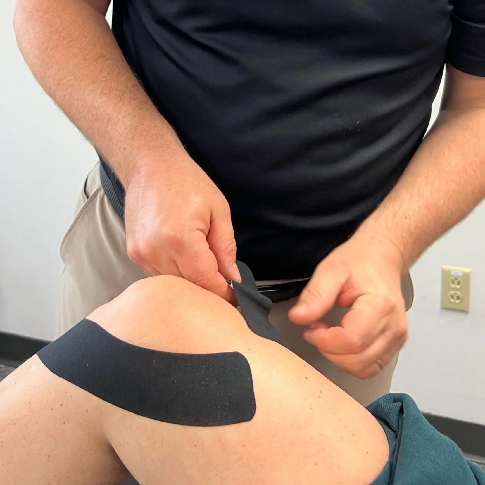 A person applying a black kinesiology tape on another person's shoulder during a treatment or therapy session.