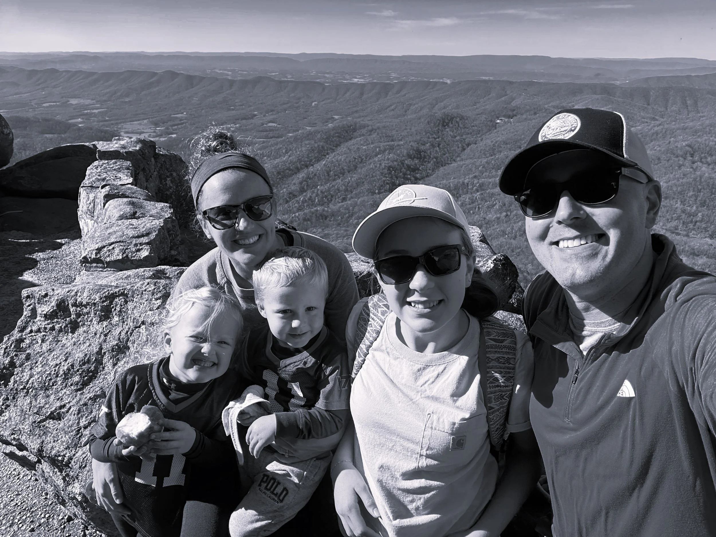 A family of five taking a selfie on a mountain summit with a panoramic view of hills and valleys in the background.