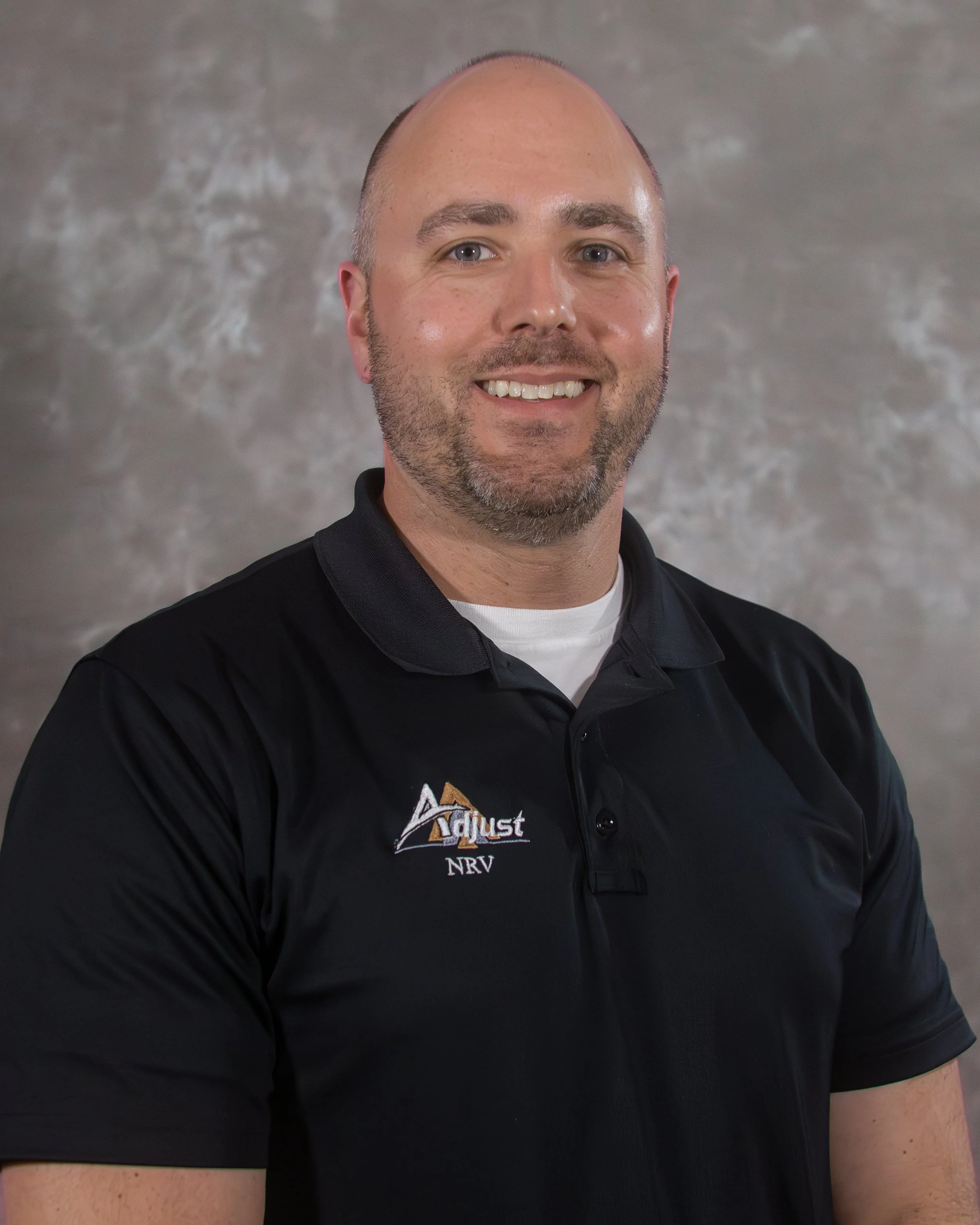 A man in a black collared shirt smiling, with a logo on the shirt that says 'Adjust NRV'.