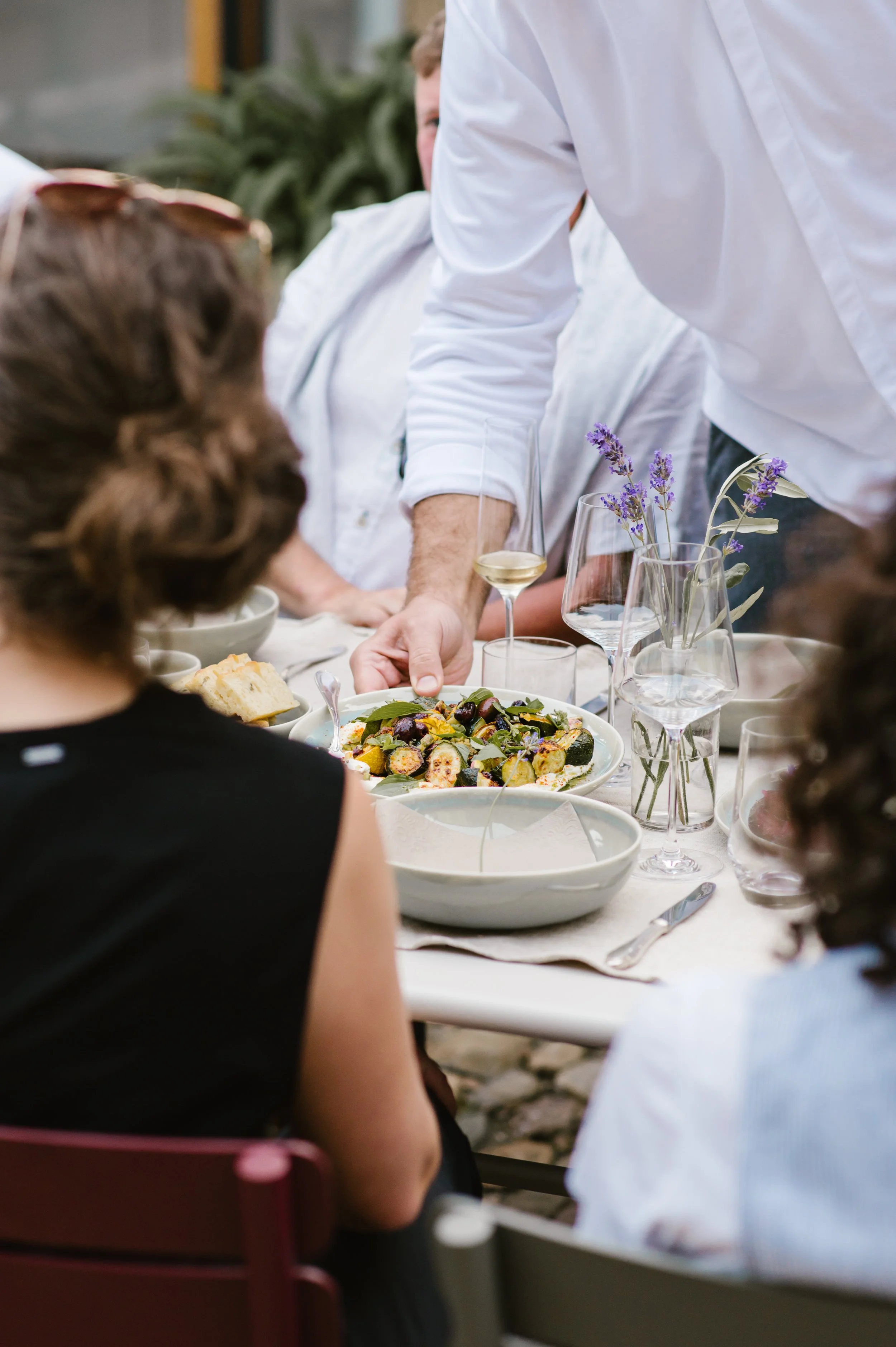 Menschen beim Servieren eines Tellers bei einem geselligen Essen, mit Weingläsern und Blumen auf dem Tisch.