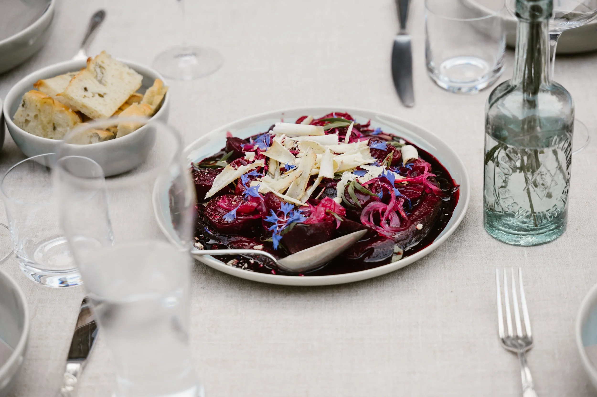 Bunter Salat mit Roten Beeten, Zwiebeln, Sprossen und Käse auf einem weißen Teller, umgeben von Wasserflaschen, Gläsern, Besteck und einem Brotkorb auf einem gedeckten Tisch.