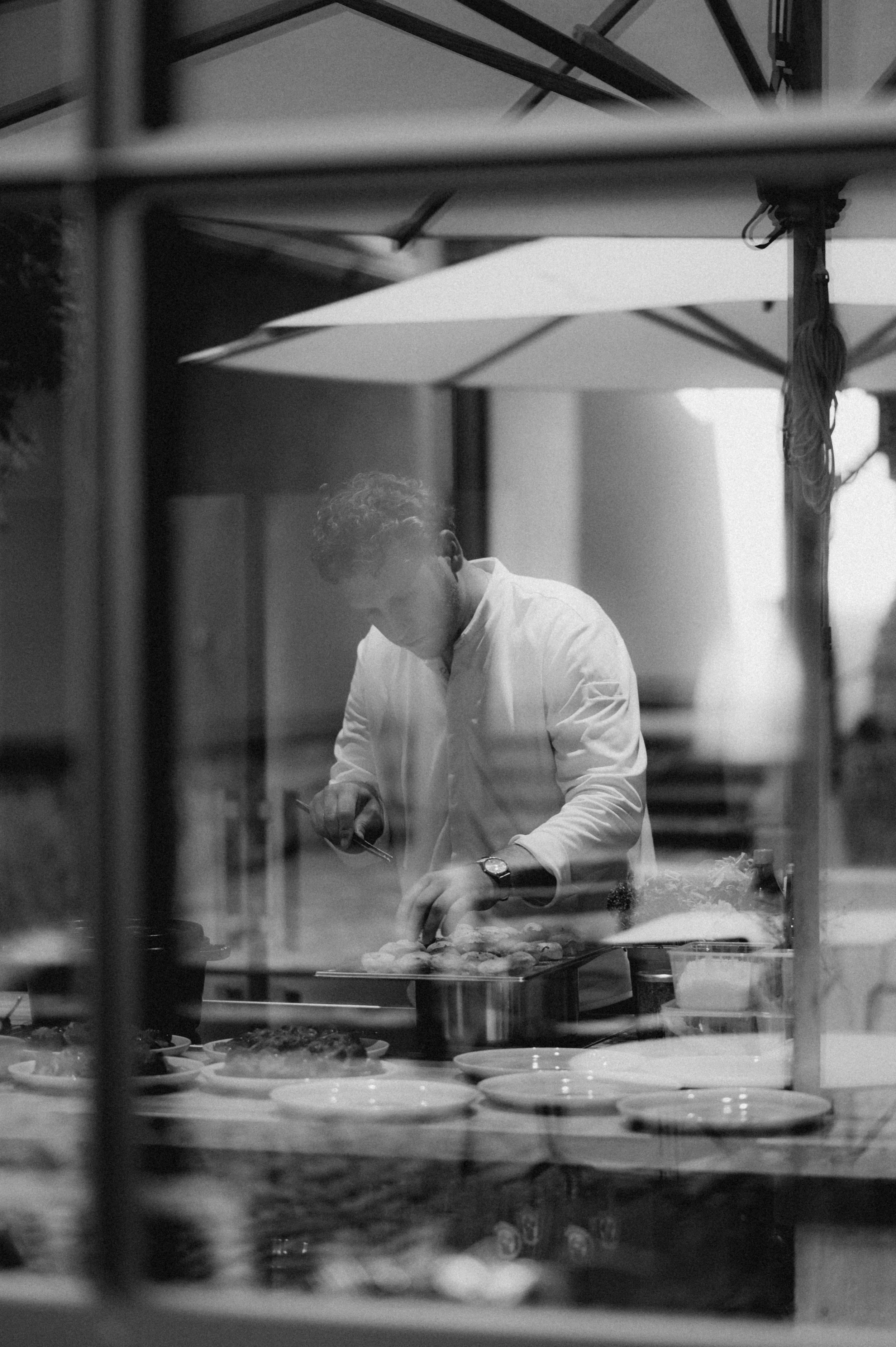 Ein Koch bereitet Speisen in einer Küche vor, gesehen durch eine Glasscheibe in Schwarzweiß, mit Tischen und Küchenutensilien im Hintergrund.