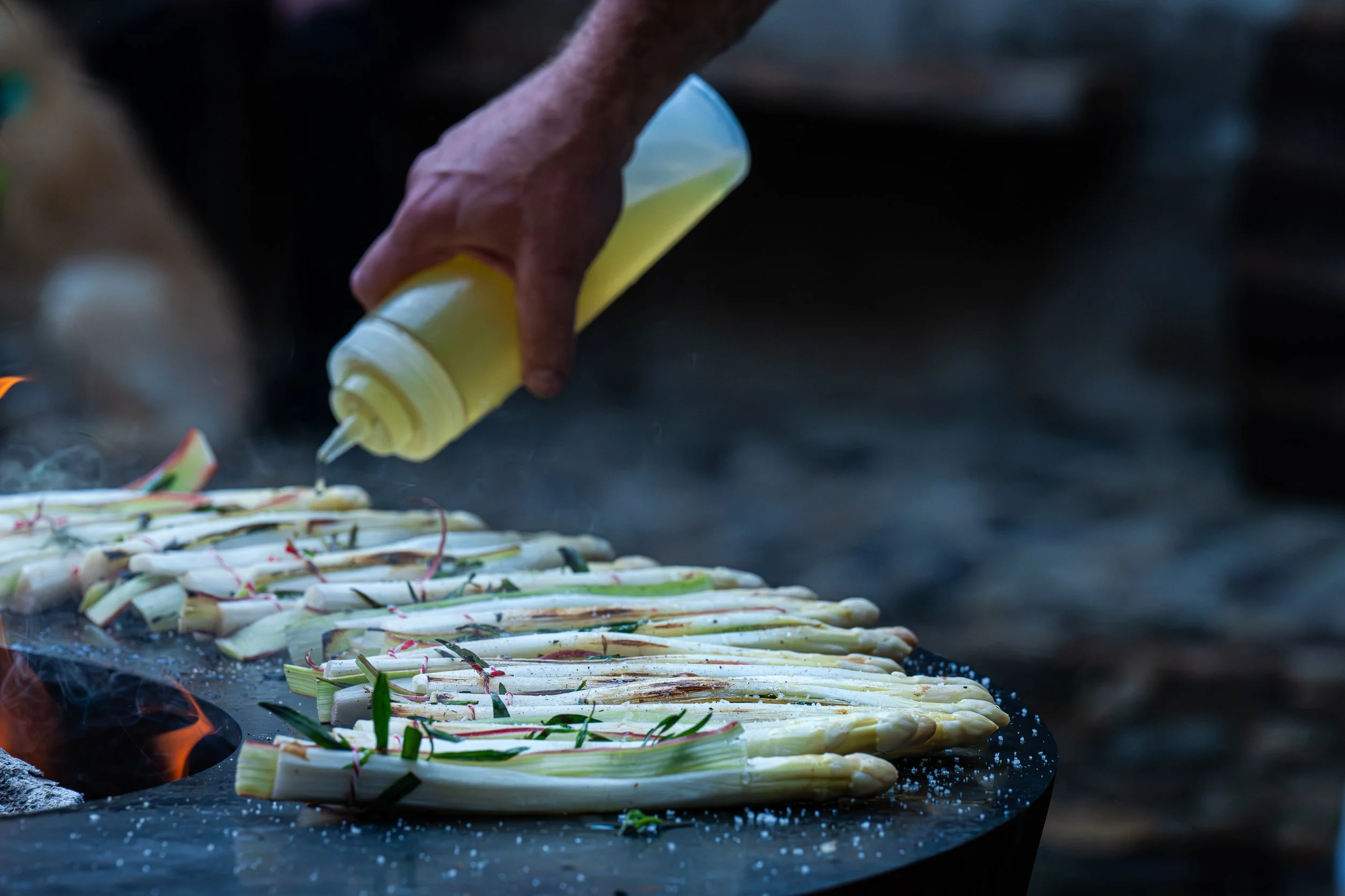 Hand mit Käsespäne auf einer heißen Grillfläche, wobei der Käse schmilzt. Es sind mehrere Stücke Käse, die mit grünen Kräutern und roten Bändern zusammengebunden sind, während sie auf dem Grill sind.