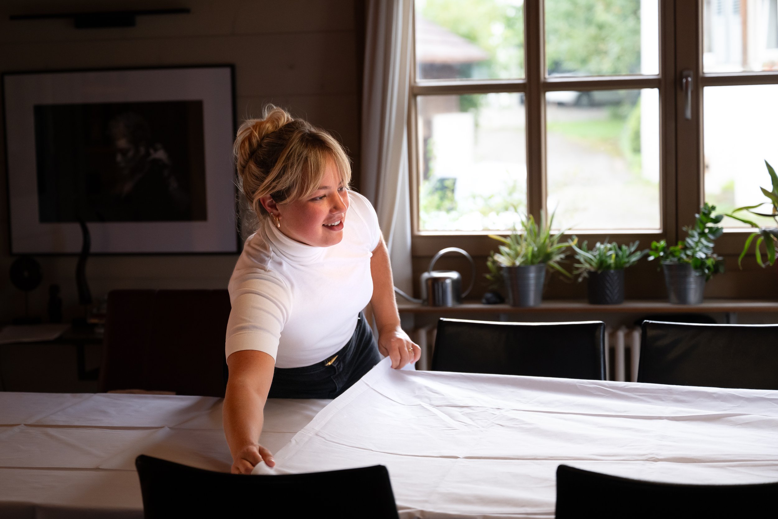 Frau beim Decken eines Tisches in einem hellen, gemütlichen Raum mit Pflanzen auf der Fensterbank.