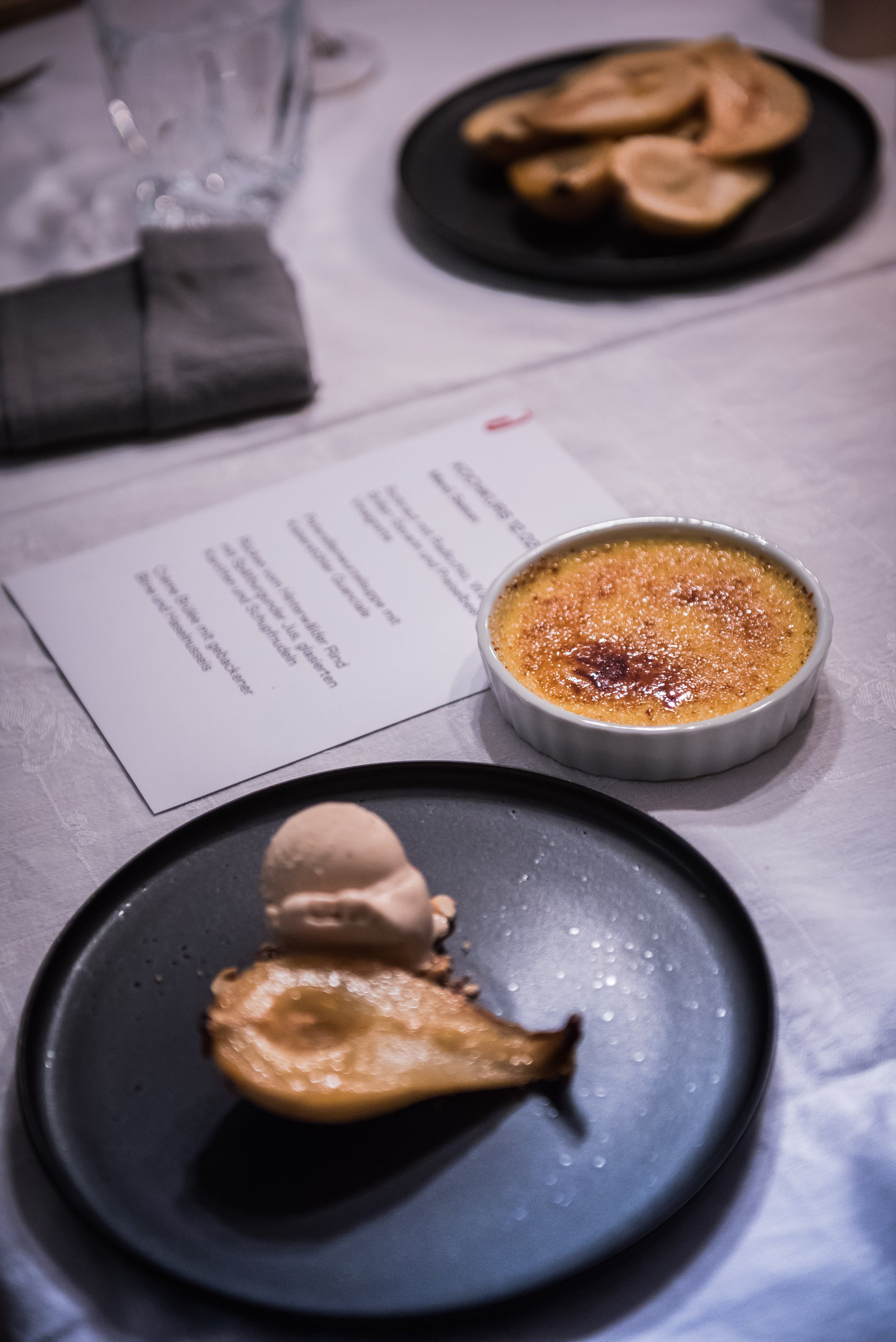 Ein Dessert mit gebratener Birne, einer Kugel Eis und einer Creme brûlée auf einem schwarzen Teller, mit einer Speisekarte und einem Glas Wasser im Hintergrund.