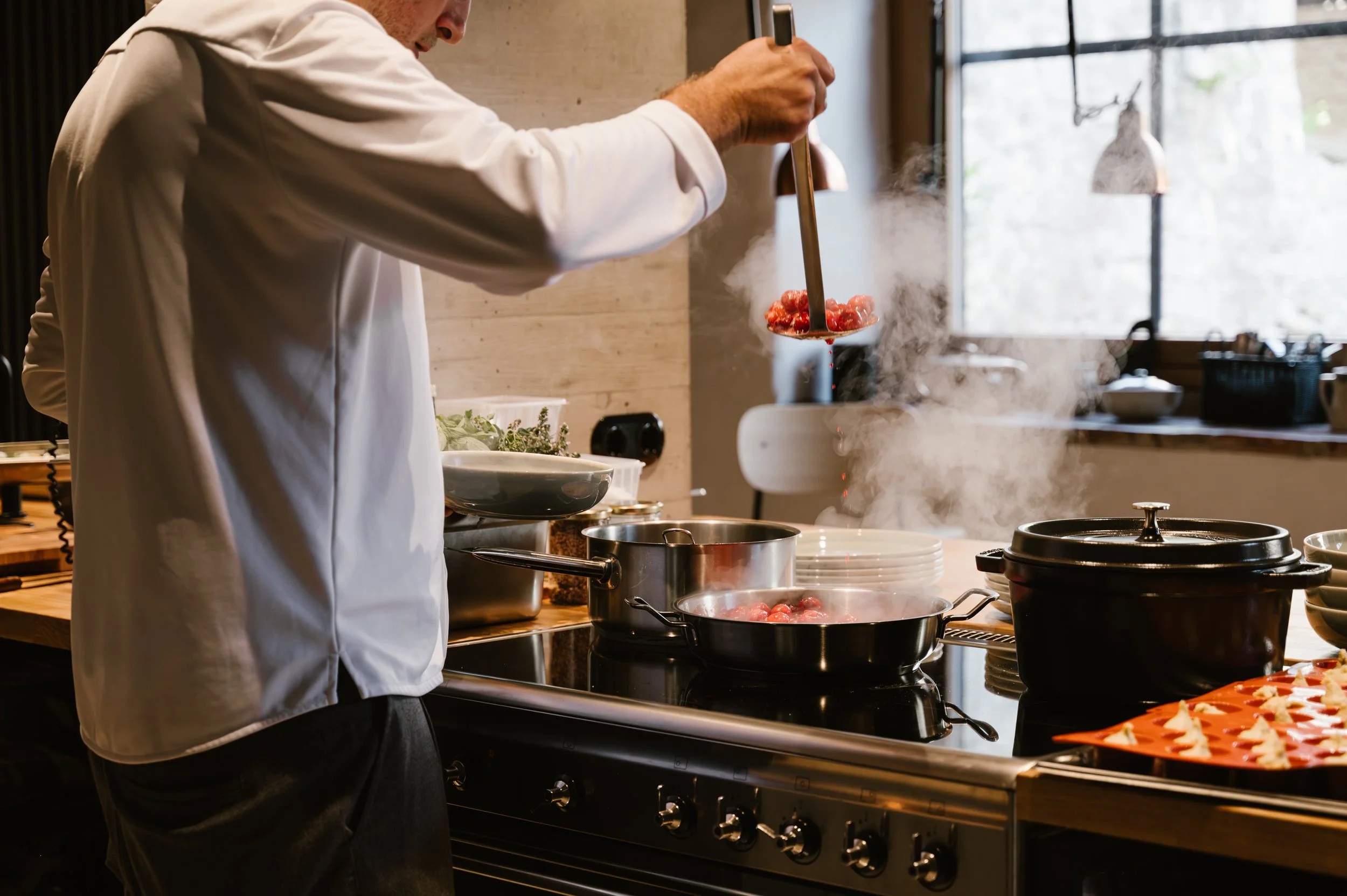 Ein Koch in Weiß steht vor einem Herd und kocht, während er eine Pfanne mit Tomaten hochhält. Es dampft und es gibt verschiedene Kochutensilien und Geschirr auf der Küchenarbeitsplatte, mit einemfenster im Hintergrund.