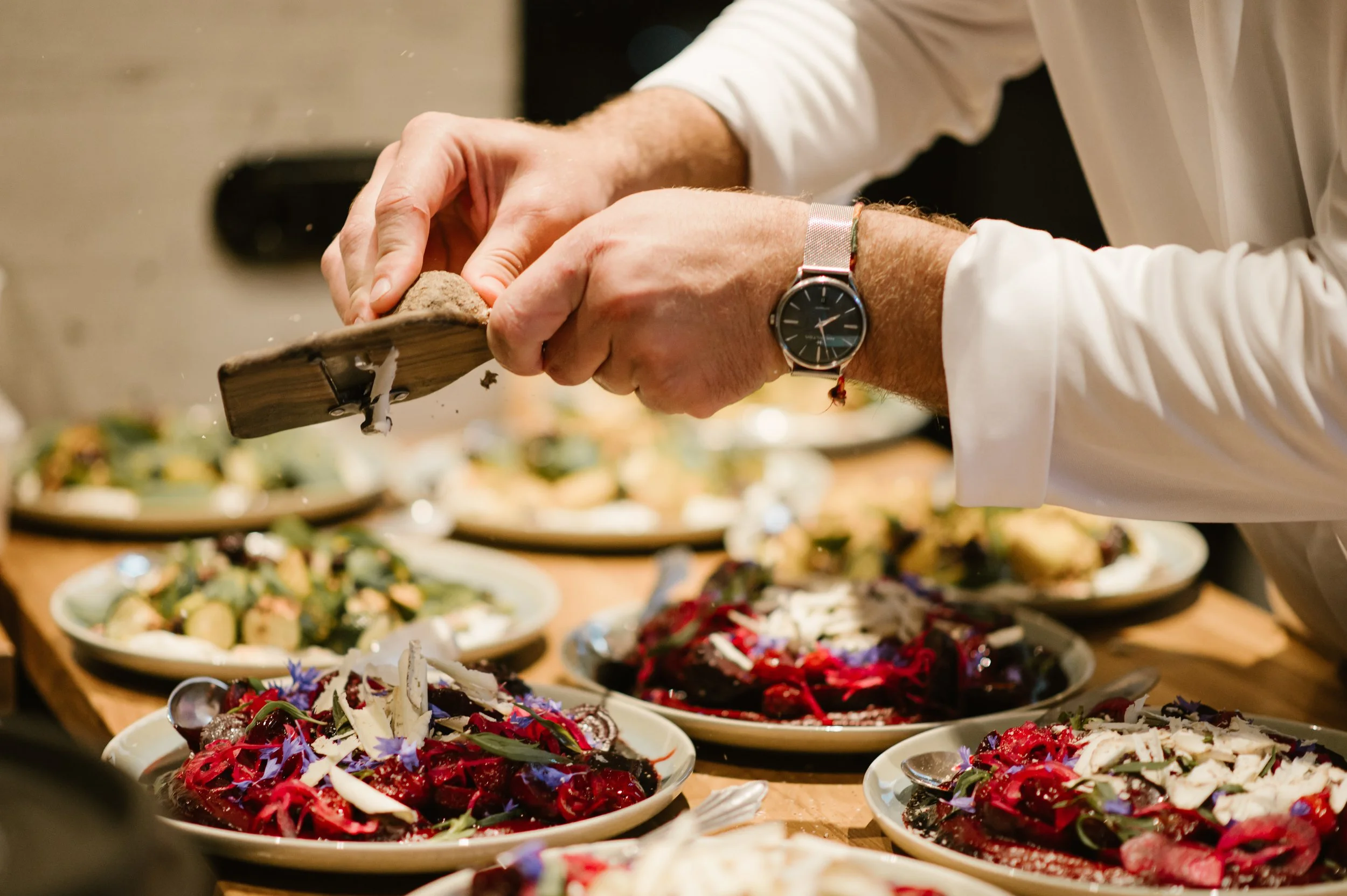 Ein Koch streut Gewürz oder Salz auf Gerichte, die auf einem Tisch mit mehreren Platten serviert werden, während im Hintergrund weitere Speisen sichtbar sind.