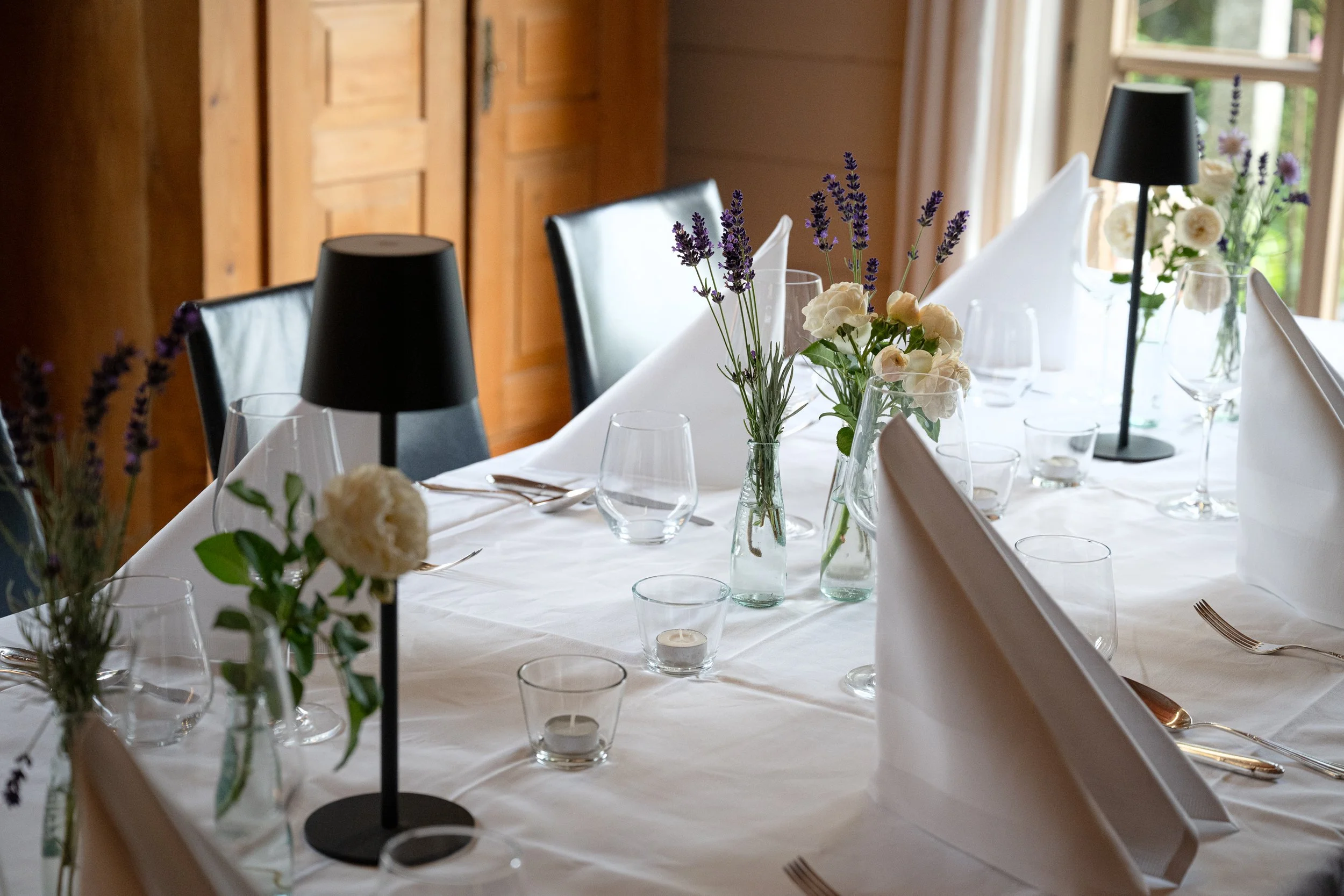 Elegant gedeckter Esstisch mit weißen Napkins, Gläsern, kleinen Kerzengläsern, Lichtern, Lavendel- und weißen Blumen in Glasvasen, schwarze Tischlampen, in einem Holzhaus mit großen Fenstern.