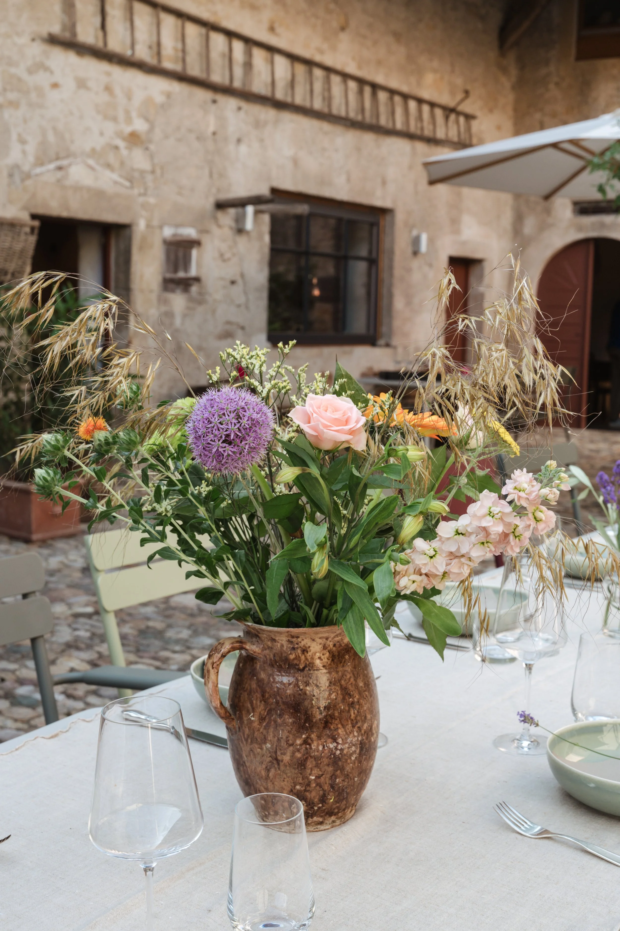 Blumenstrauß in einer braunen Keramikvase auf einem festlichen Tisch, im Hintergrund eine alte Steinmauer eines Gebäudes.