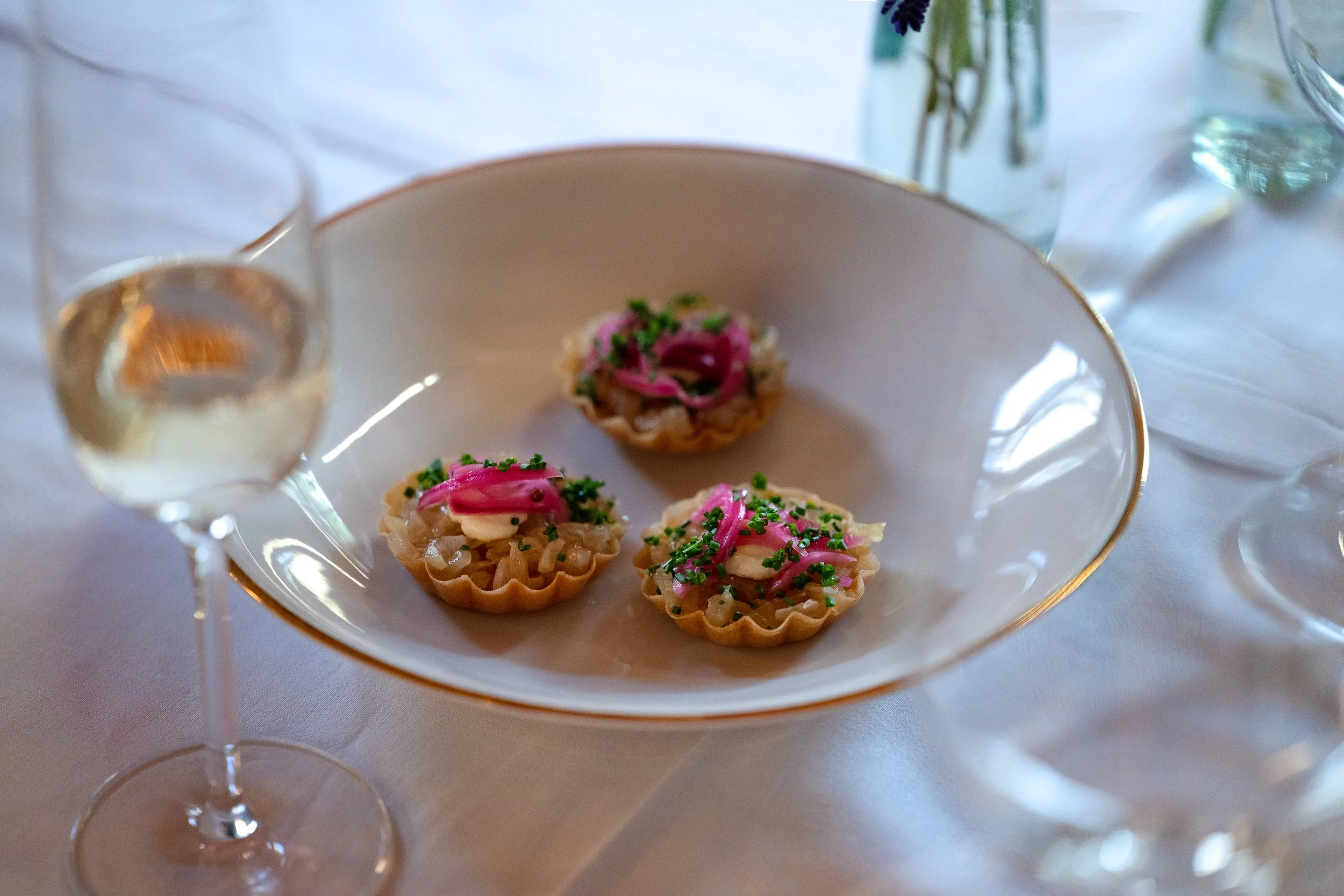 Drei kleine Tartlets mit rosa Zwiebeln und Schnittlauch auf einem weißen Teller, umgeben von Weingläsern auf einem gedeckten Tisch.