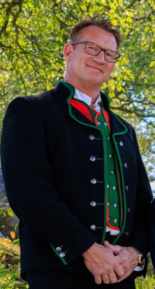 En mann i tradisjonell, fargerik drakt står utendørs foran grønne trær, smilende og ser mot kamera.