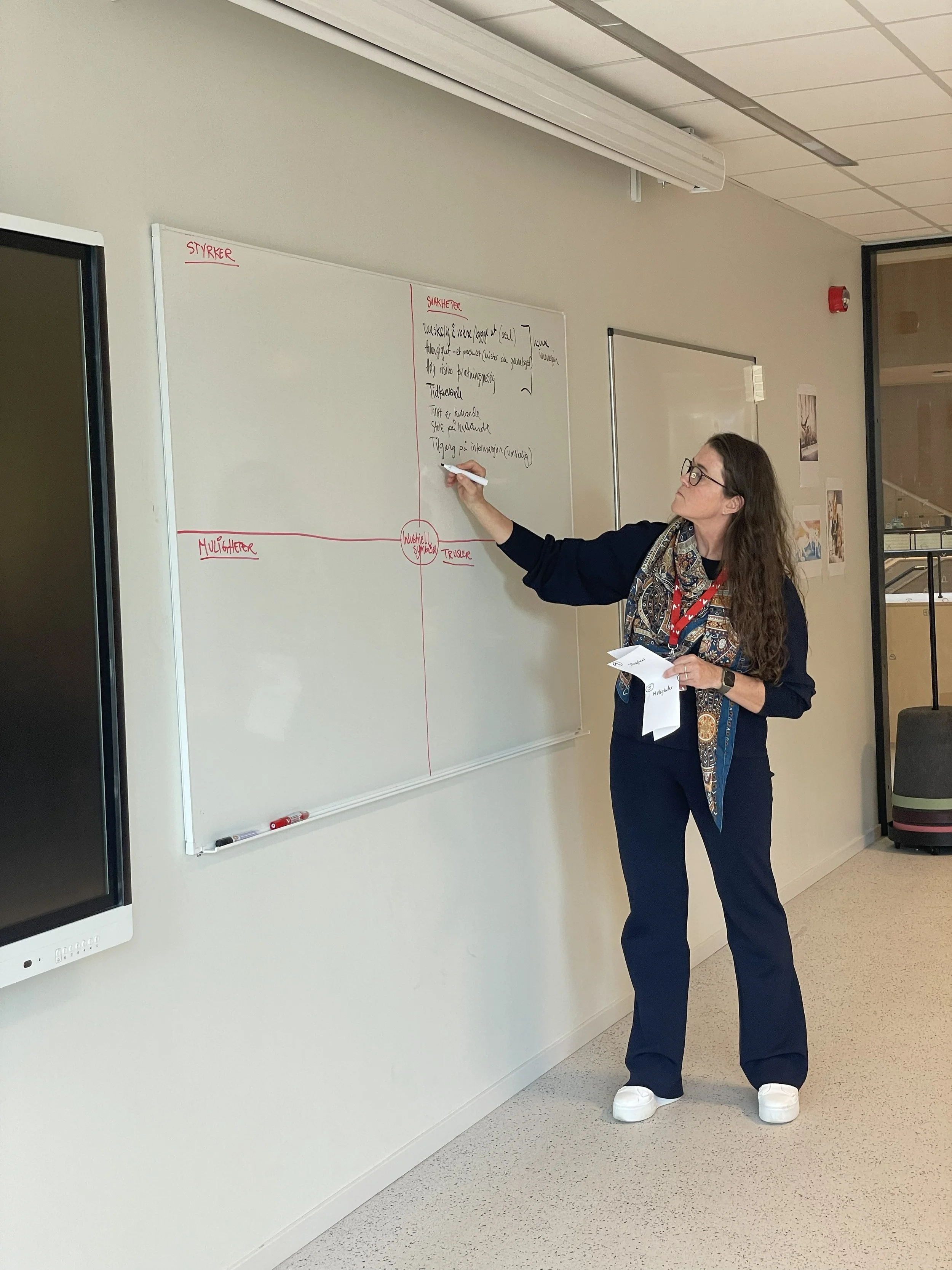 En kvinnelig person står foran en whiteboard og skriver med svart penn. Personen har briller, langt bølget hår, og er iført en mørk blazer, skjorte med mønster, og store hvite sneakers. Whiteboardet har røde overskrifter og svart skrift, og personen holder en papir i hånden.