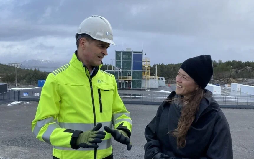 To personer snakker ute på et industriområde, en i en høy-vis jernbanedrakt med hjelm, den andre i mørk jakke og lue, med fabrikkanlegg i bakgrunnen.