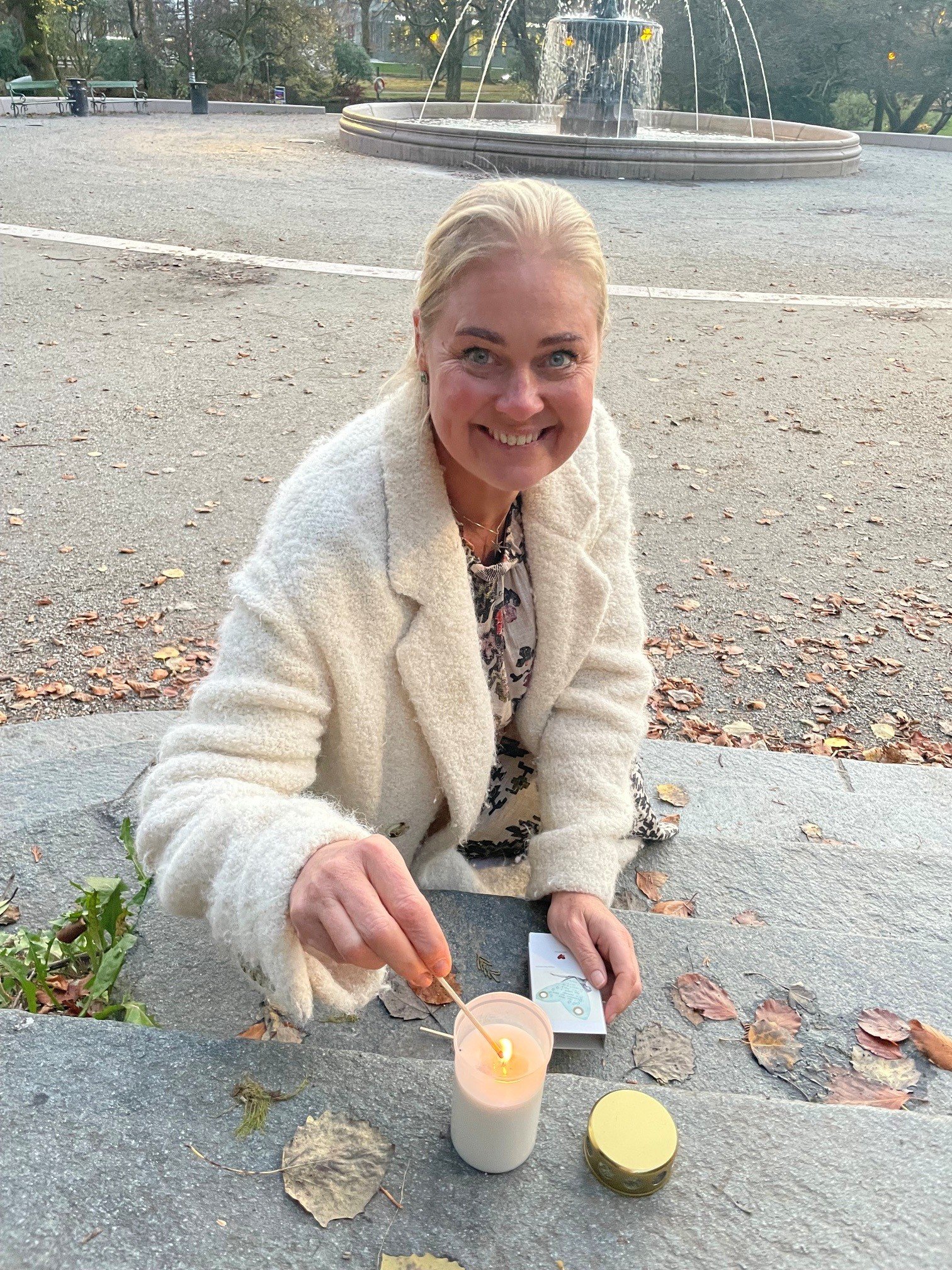 Et smilende eldre kvinne med blondt hår i en lys jakke som holder en tynn pinne over en lysende stearinlys på et benkeppe i en park, omgitt av tørre løv på bakken.