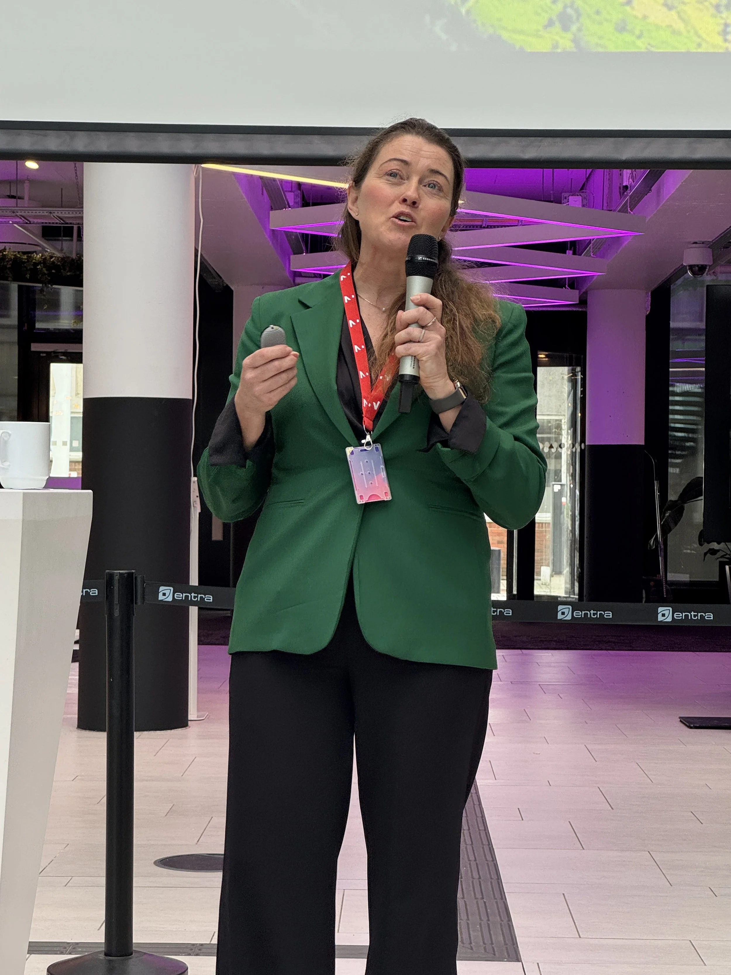 En kvinne i grøn blazer snakker inn i mikrofon, står i et konferanserom med purple belysning i bakgrunnen.