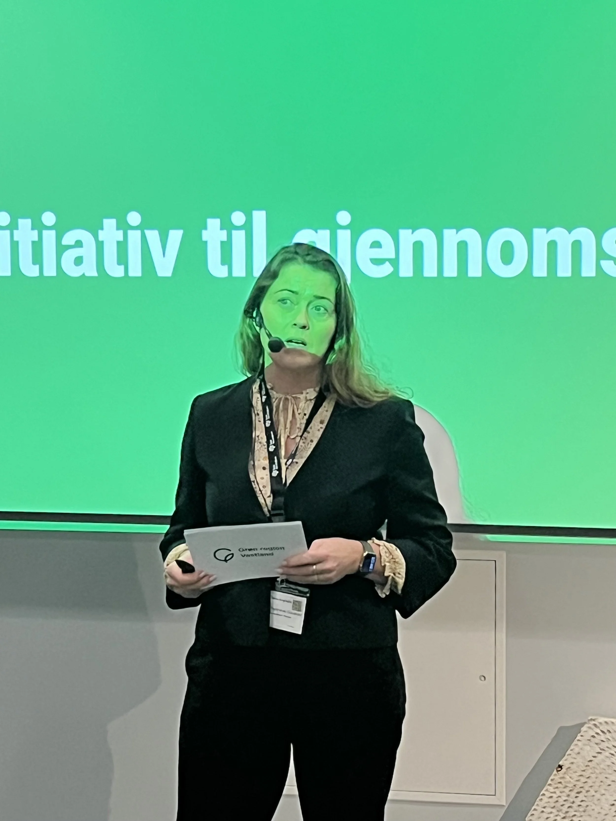 Kvinne med mikrofon, som står foran en grønn skjerm med delvis synlig tekst.