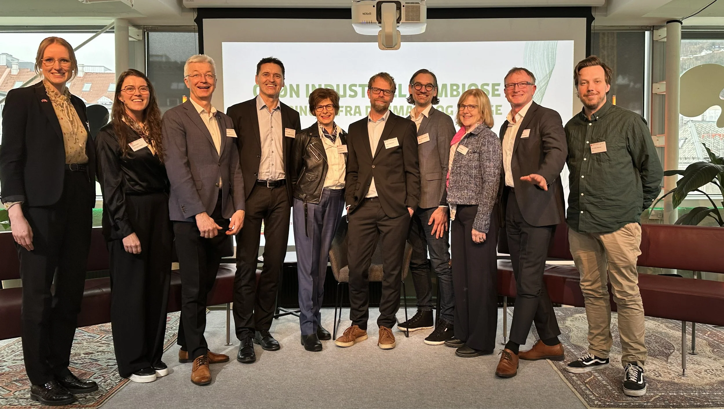En gruppe på tolv mennesker står foran en projektor og en skjerm med teksten «ON INDUSTRIAL AMBITION» i et rom med store vinduer og byutsikt.