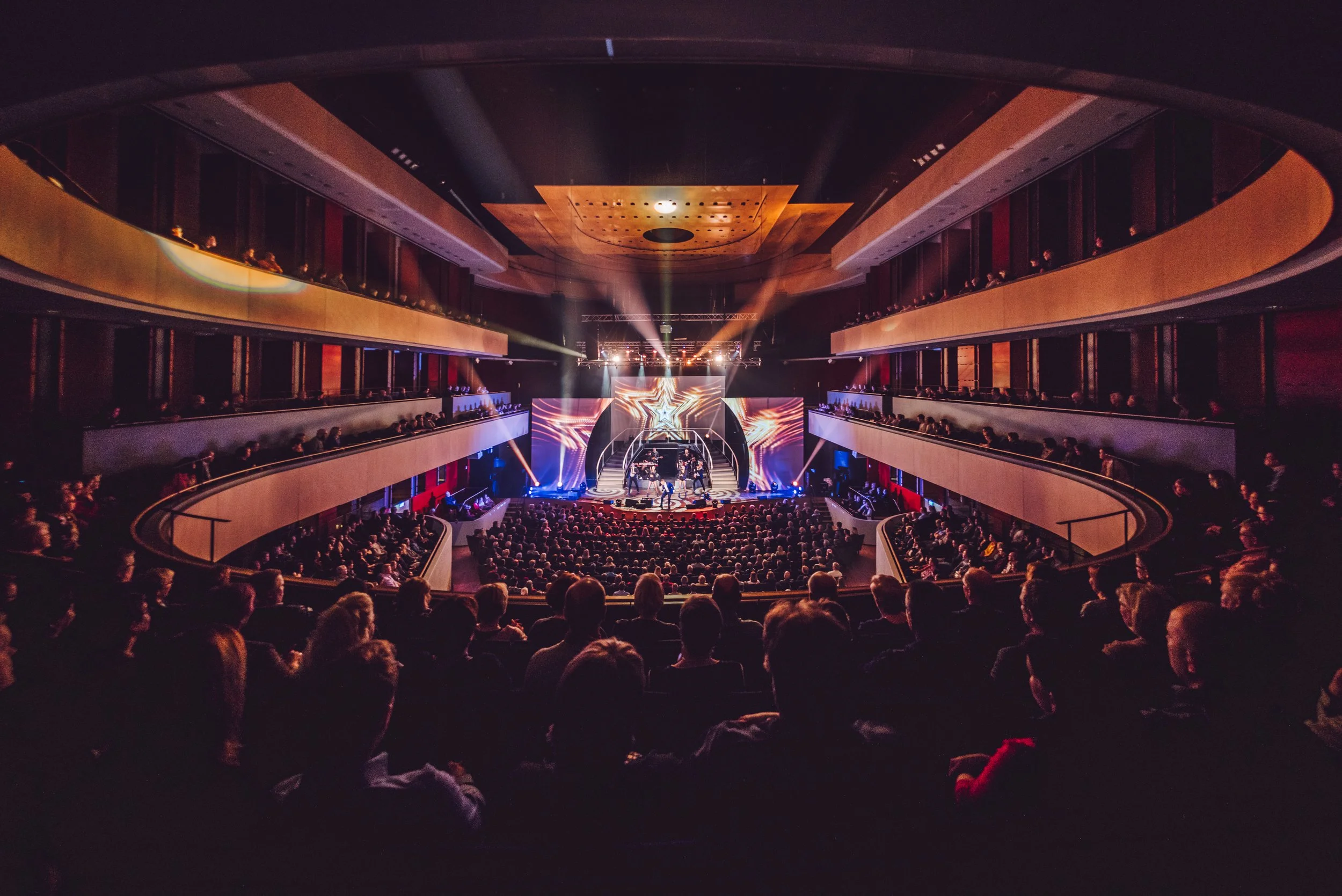 An interior view of a theater with multiple balcony levels filled with an audience watching a stage performance with bright lights and visual effects.