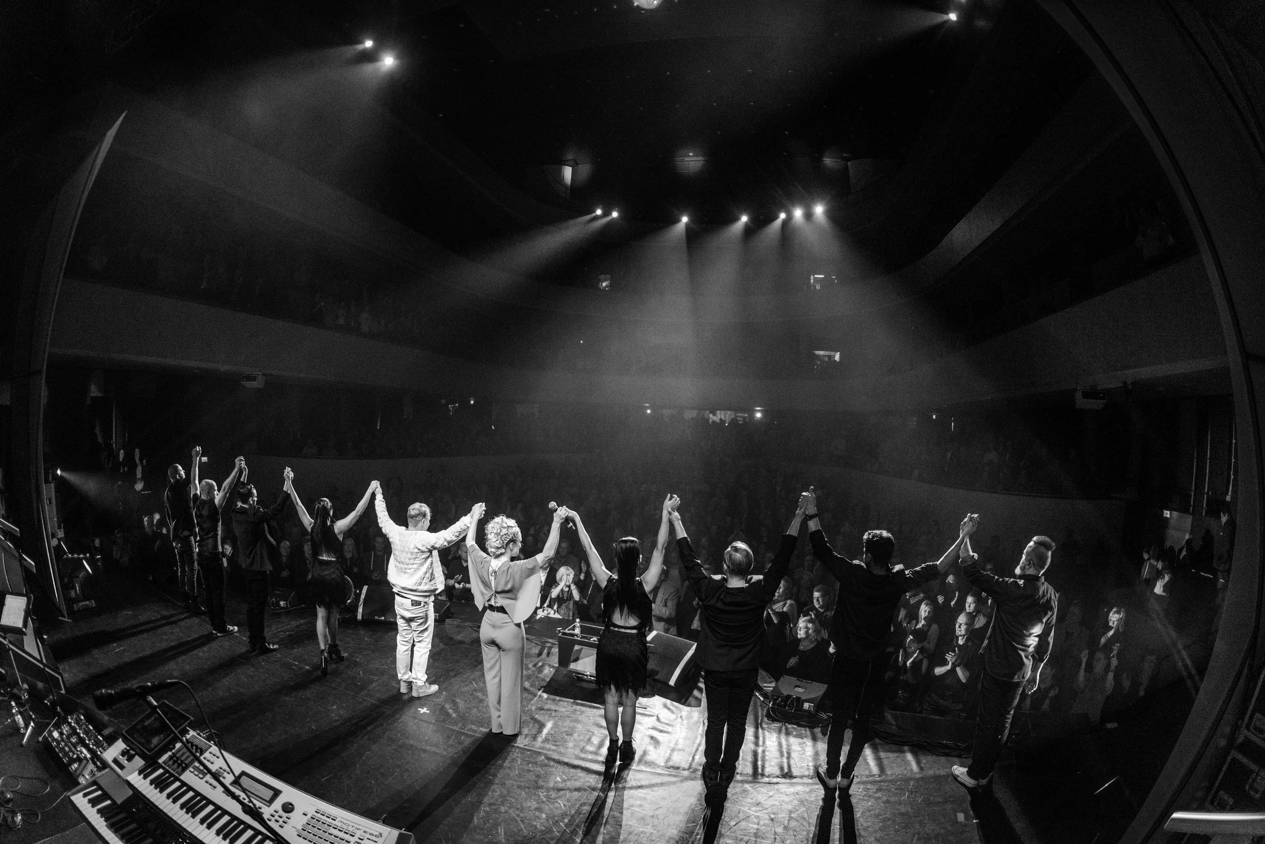 Group of performers holding hands and bowing on stage after a concert, with audience clapping in the background.