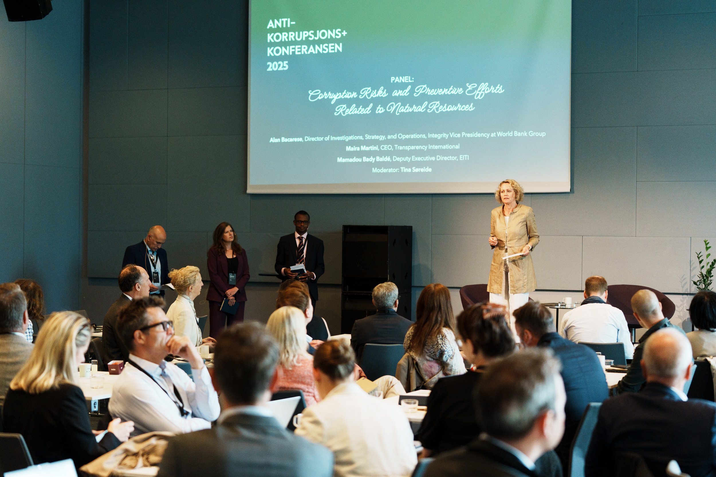 En kvinne snakker på en konferansescene foran et stort skjermbilde med teksten 'ANTI-KORRUPSJONS+KONFERANSEN 2025' og panelinformasjon, mens en gruppe mennesker sitter og hører på.