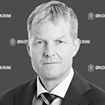 Et svart-hvitt portrett av en mann i dress og slips, med lyst hår og alvorlig uttrykk, mot en bakgrunn med logoer.